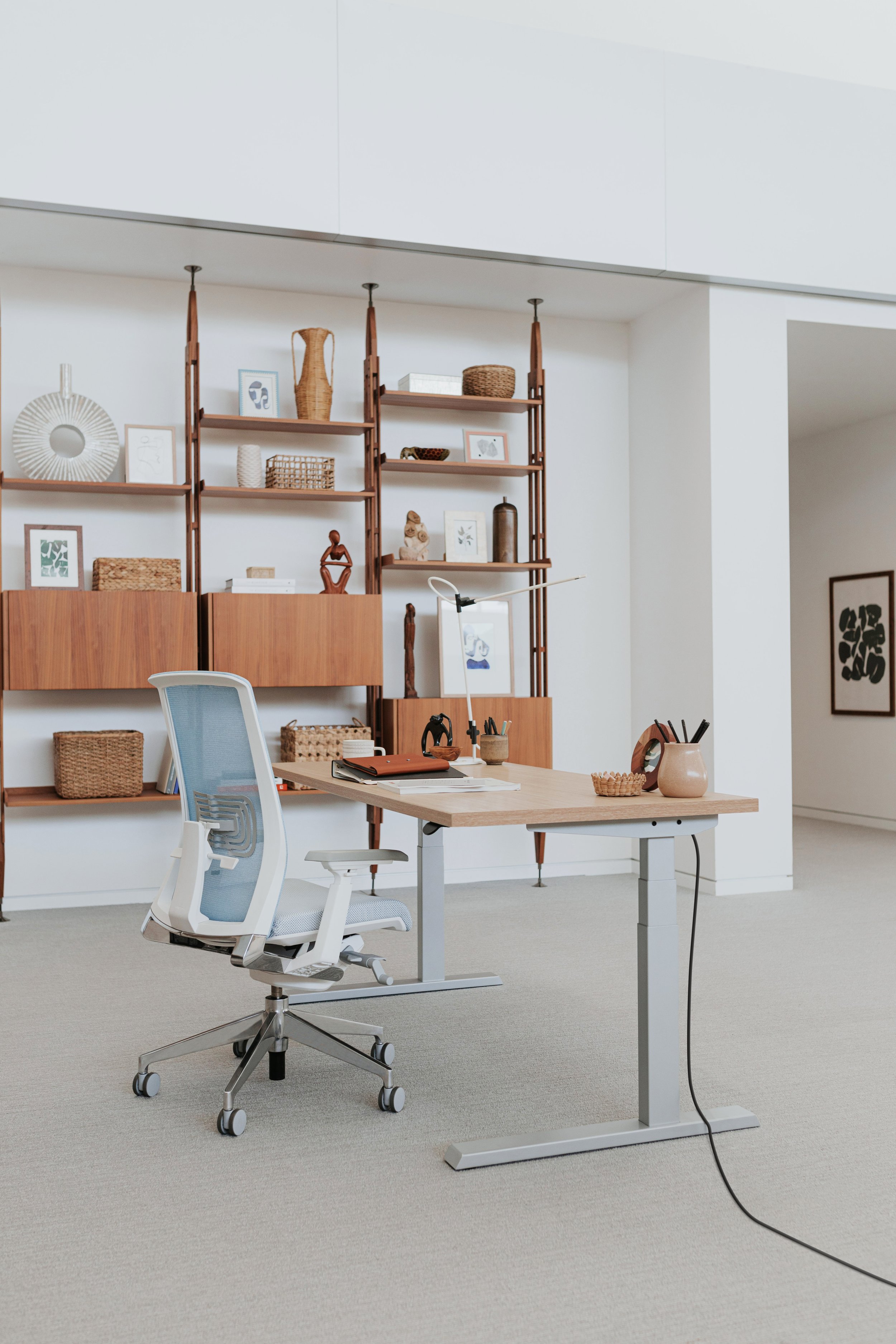 Modern home office with a wooden desk, ergonomic white chair, and decorative wooden shelving with art pieces and woven baskets.
