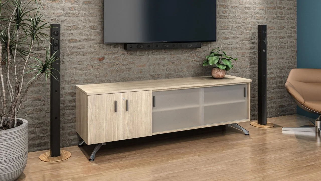 Modern living room with wooden TV stand, wall-mounted flat-screen TV, tall speakers on either side, potted plant on the stand, exposed brick wall, hardwood floor, beige armchair, and a blue accent wall.