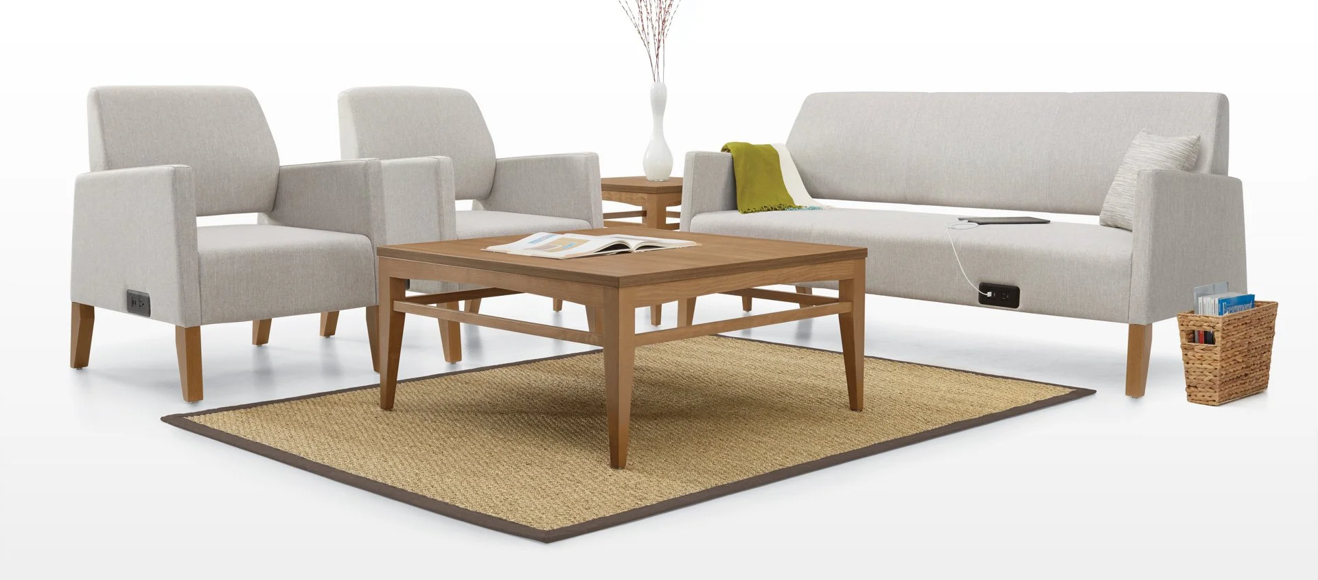 Living room with two beige armchairs, a beige sofa with a green throw blanket, a wooden coffee table with open book, a matching side table with a white vase with branches, a woven magazine basket, and a beige area rug.