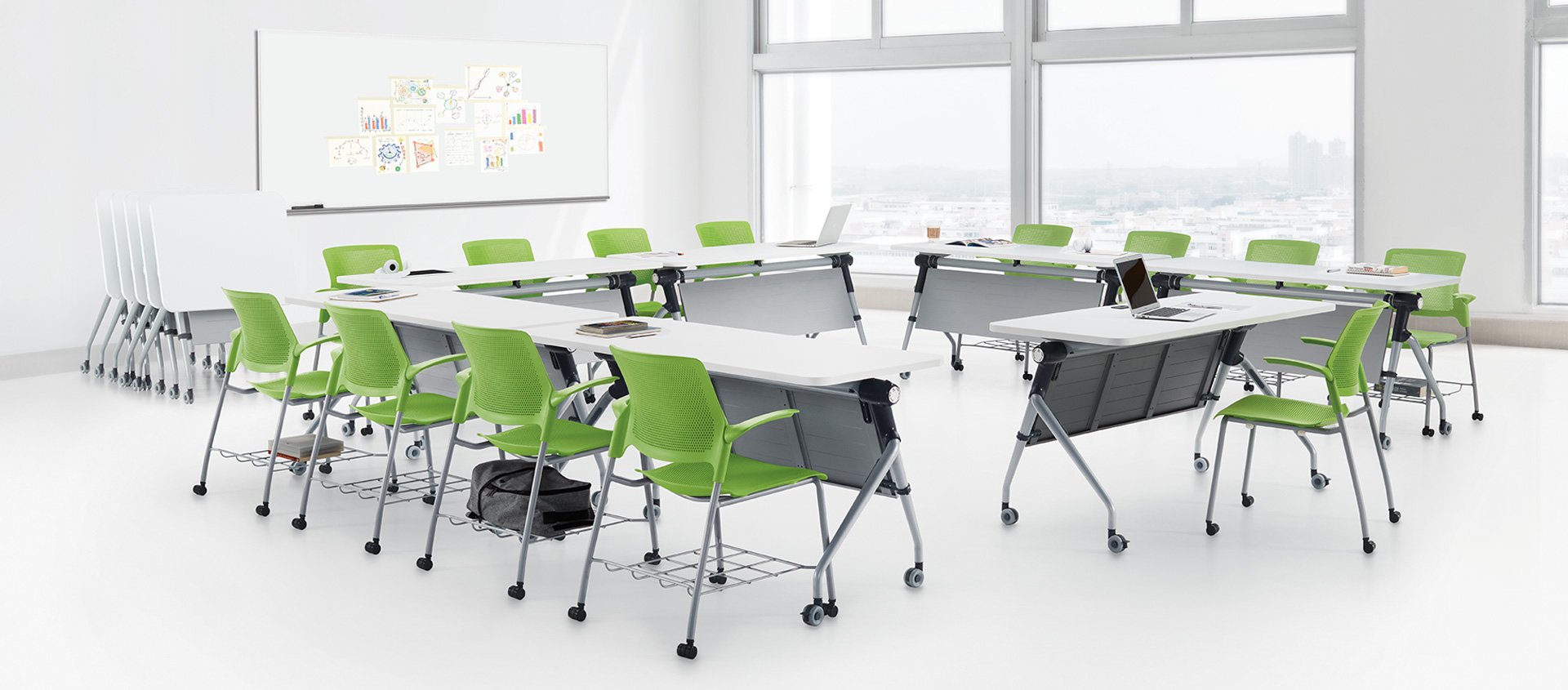 Modern classroom or conference room with white tables, green chairs, laptops, and large windows with city view.