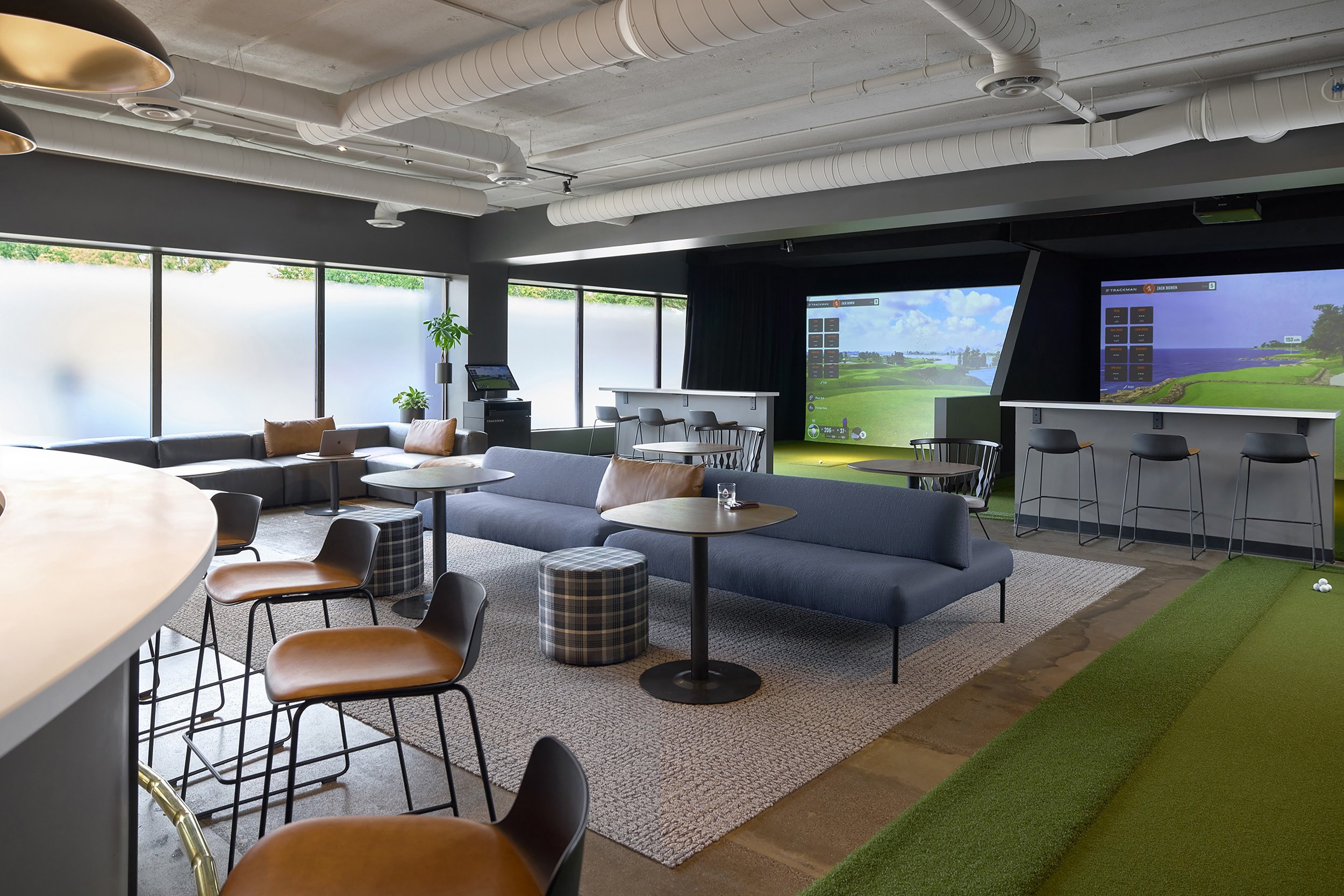 Interior of a modern golf lounge with multiple screens displaying virtual golf courses, sitting area with sofas and chairs, and a small putting green.