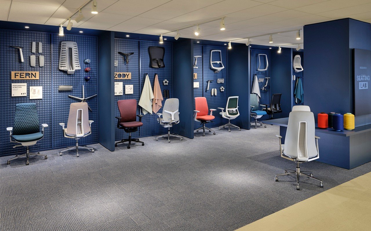 Office seating laboratory with six ergonomic chairs, various backrests and seat cushions, and fabric samples, organized by body parts, in a blue-walled room with spotlights.