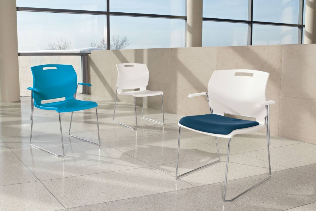 Three modern chairs with plastic seats and backs in blue and white, and metal legs, arranged in a waiting area near large window panels with a view of trees.