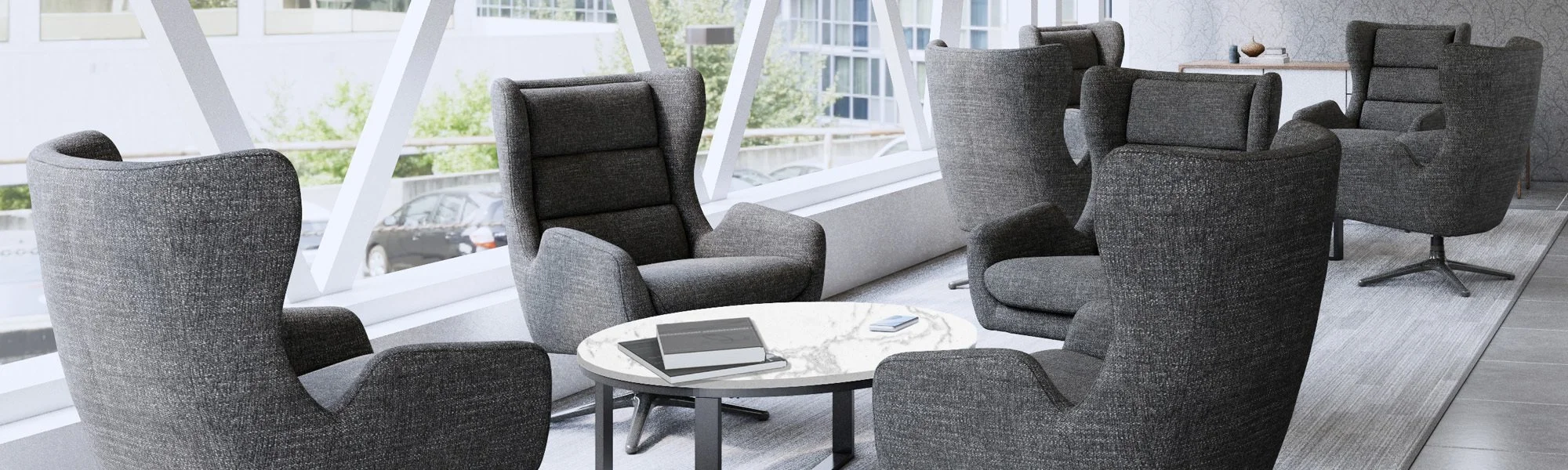 A modern office lounge area with dark grey chairs around a round marble coffee table near large windows, with a view of parked cars outside.