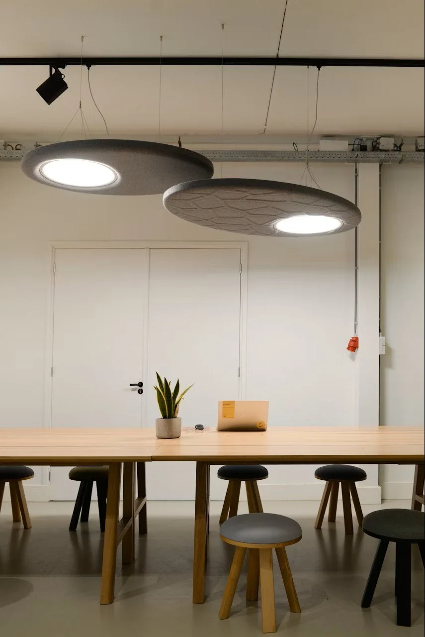 Modern conference room with a large wooden table, six stools, potted plant, and a laptop. Ceiling-mounted lighting fixtures and a door are visible in the background.