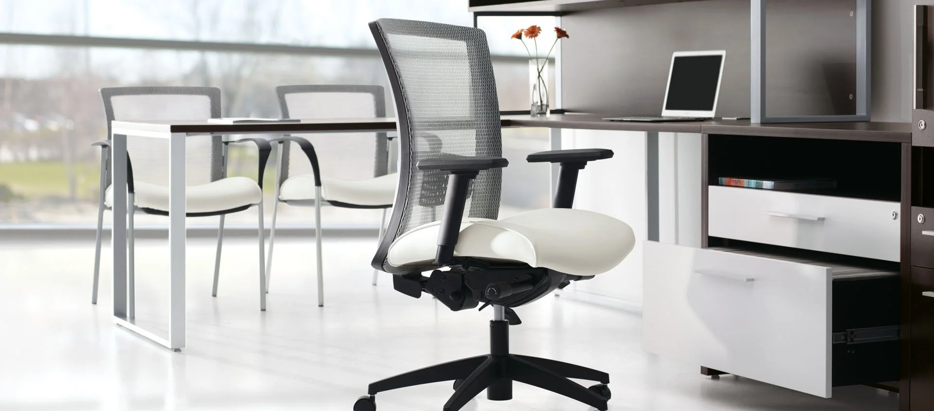 Modern office space with a black and white ergonomic office chair, white chairs around a table, and a wooden desk with a laptop, a vase with orange flowers, and storage drawers under a large window.