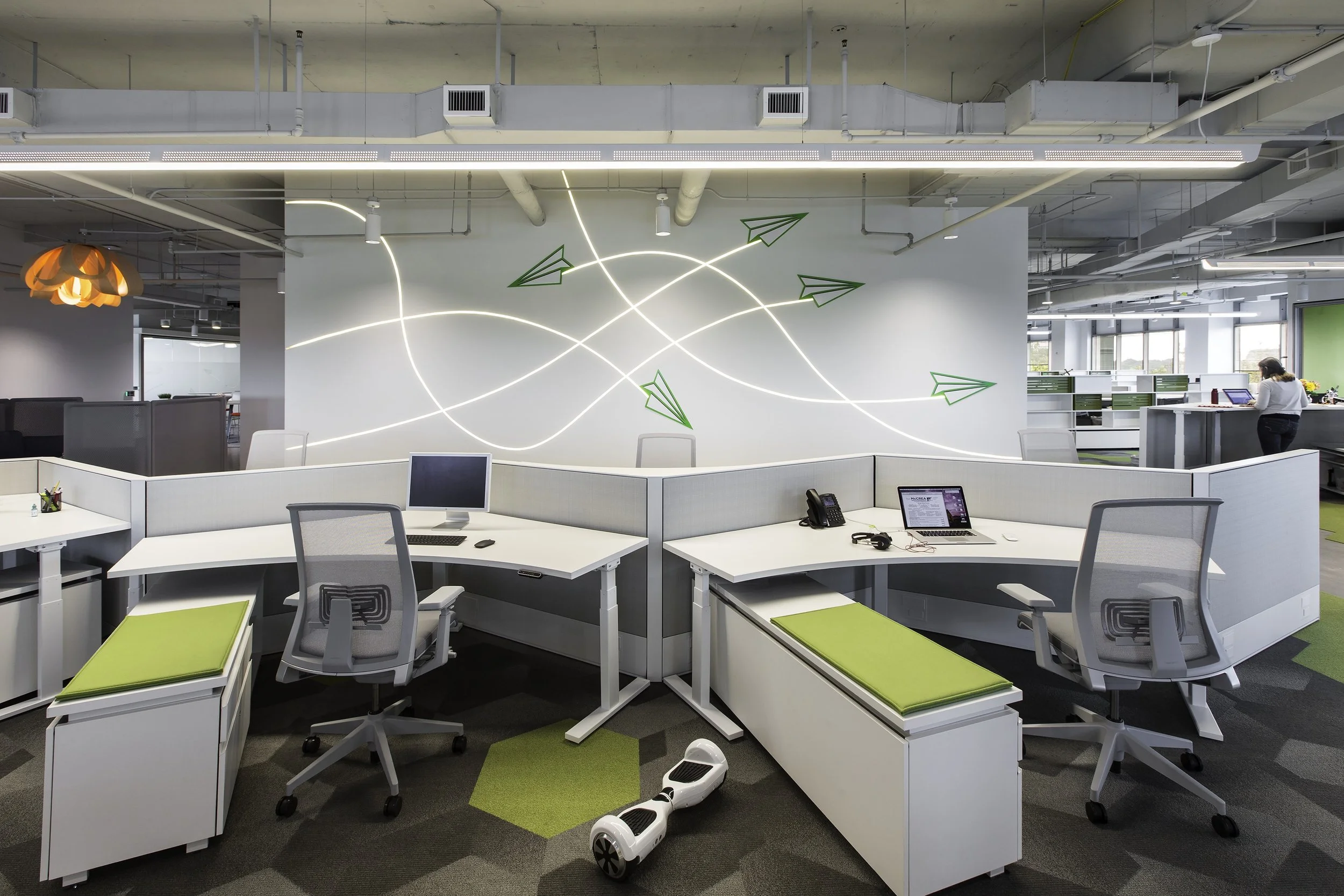 Modern office space with white desks, gray chairs, a hoverboard on the floor, and a wall decoration with neon lights and paper airplane designs.