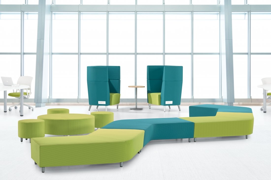 Modern office lounge with green and blue upholstered seating, high-backed chairs, and a small round table in front of large floor-to-ceiling windows.