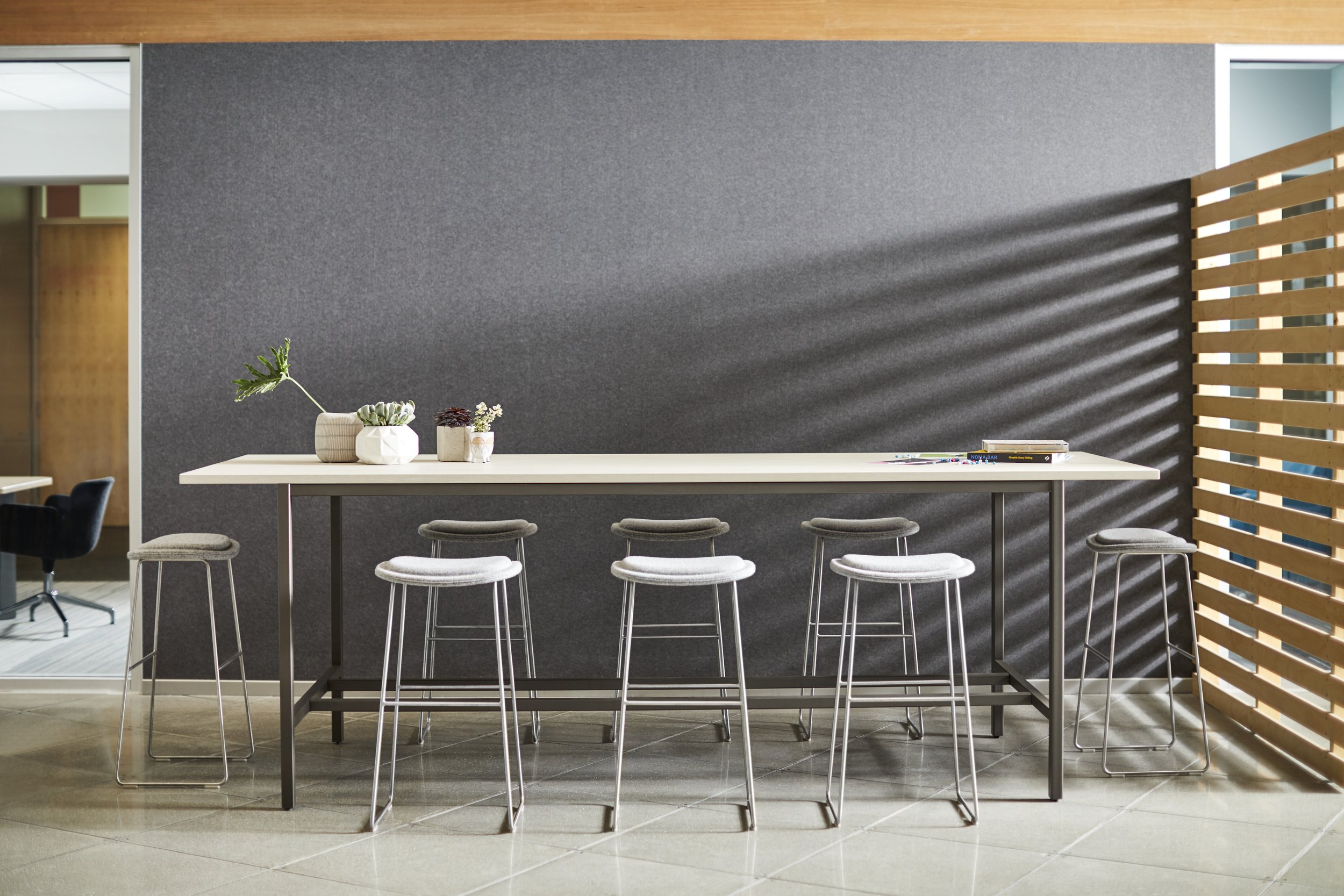 Modern office break area with a long white table, six stools, potted plants, and a gray wall with shadows, adjacent to a wooden slat partition.