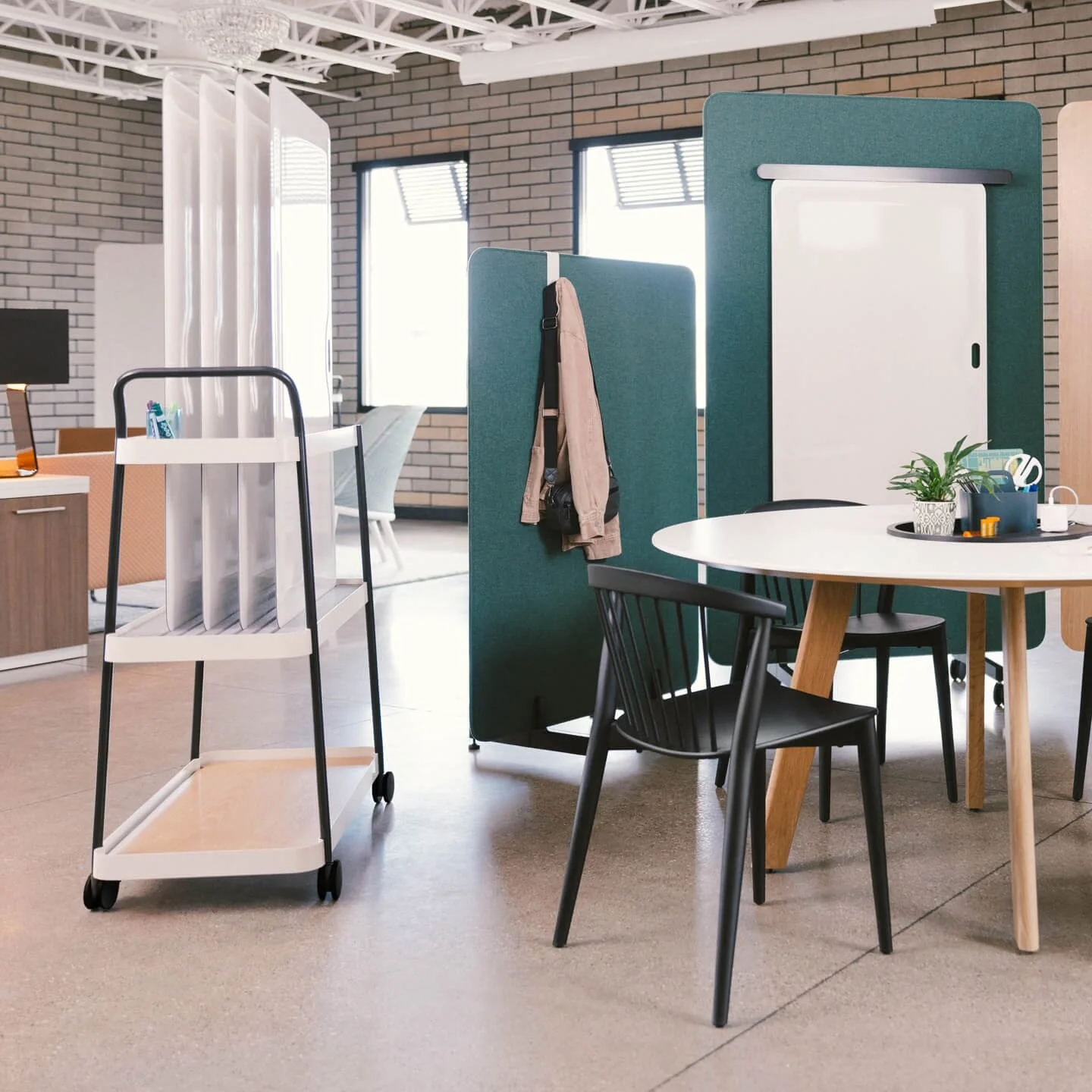 Office space with desk, black chair, potted plant, whiteboard, and privacy screens, with windows letting in natural light.