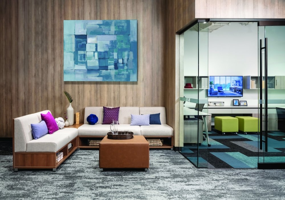 Modern office lounge area with white sectional sofa, colorful throw pillows, wooden coffee table, and an abstract blue painting on wooden-paneled wall, view into a bright enclosed office space with glass walls and green ottomans.