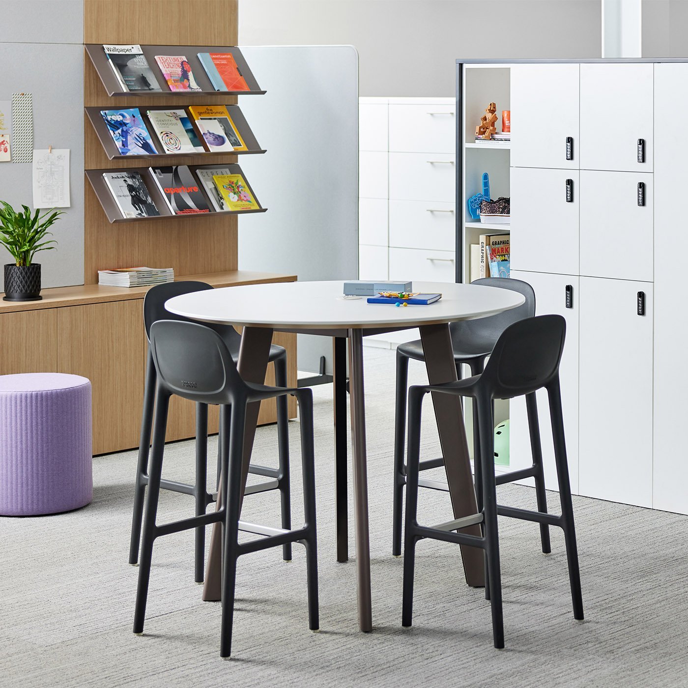 Modern office break room with a round white table, four black chairs, a purple ottoman, storage cabinets, a plant, and a metal magazine rack filled with magazines.