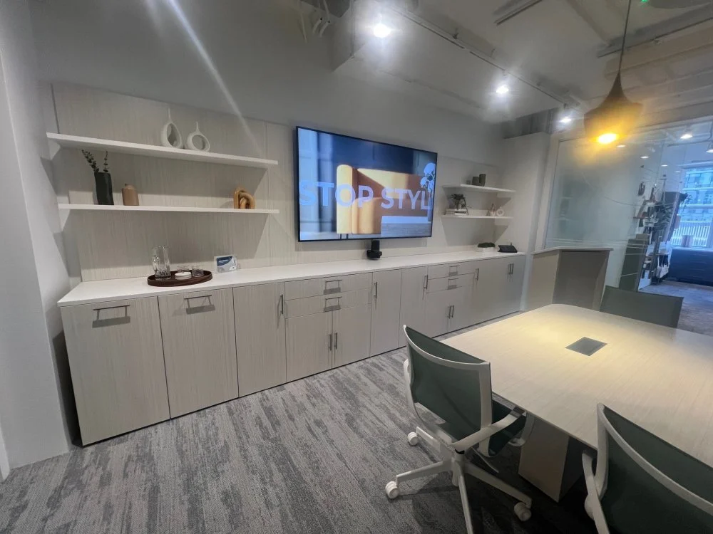 Modern conference room with a large wall-mounted TV displaying a presentation that says "STOP STYLE," a long white cabinet with decor items, and a rectangular table with green chairs.