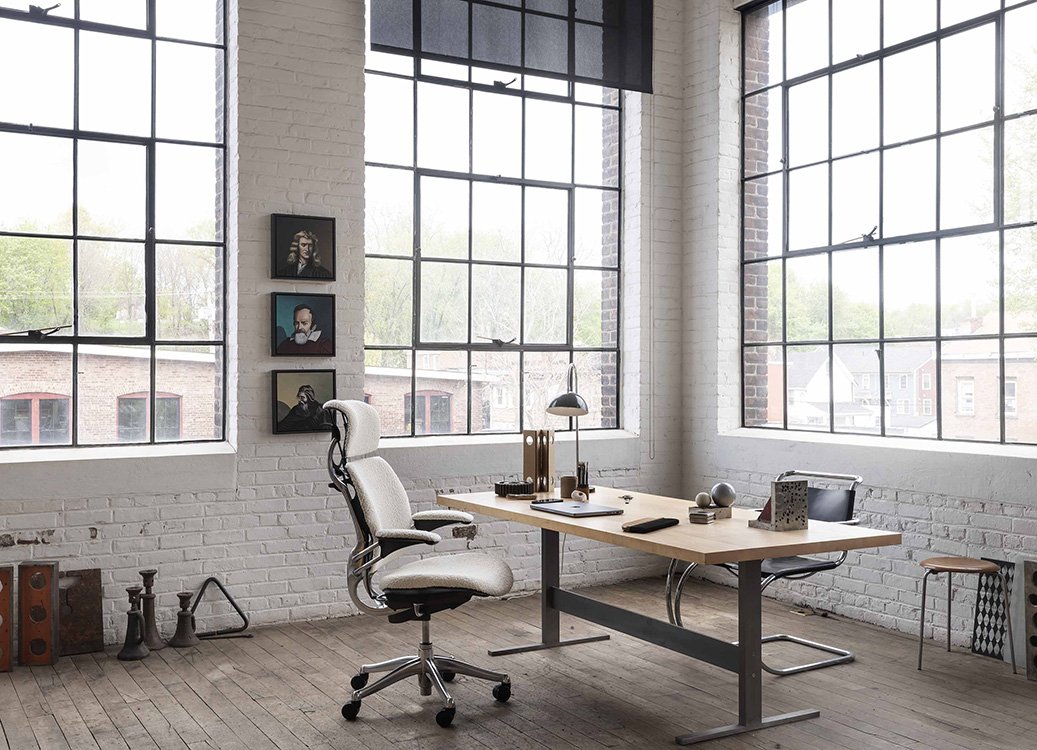 Modern office space with large windows, white brick walls, a white ergonomic office chair, a wooden desk with various office supplies, and framed artwork on the wall.