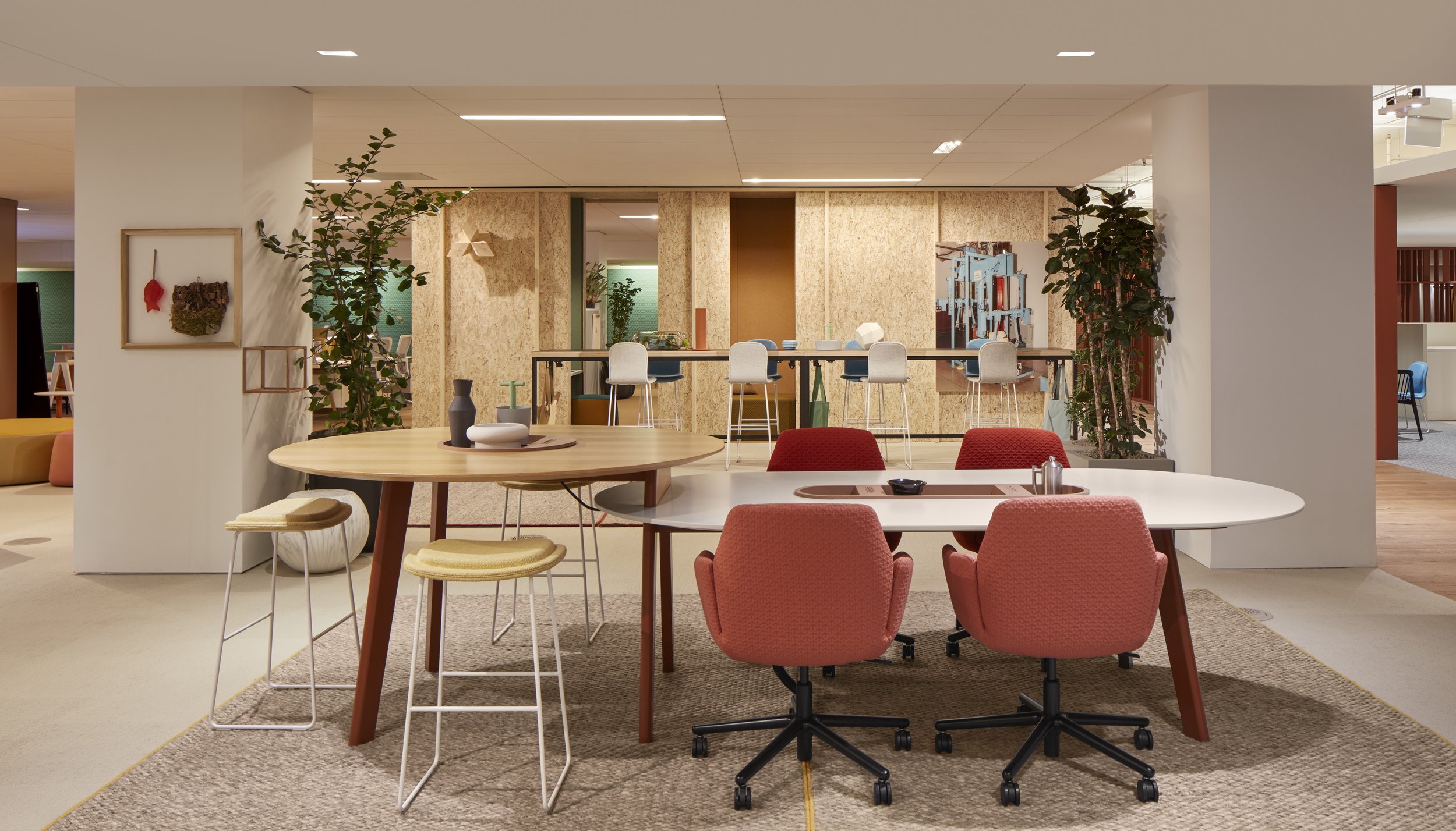 Modern open-concept office or communal workspace with round and rectangular tables, pink swivel chairs, bar stools, indoor plants, artwork, and shelving in a bright, stylish setting.