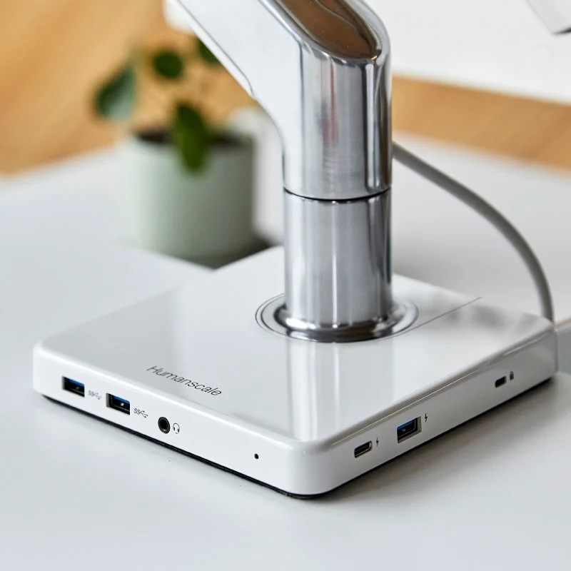 A white charging station with multiple USB ports and a power cord, with a metallic faucet extending from the top, on a white surface with a blurred potted plant in the background.