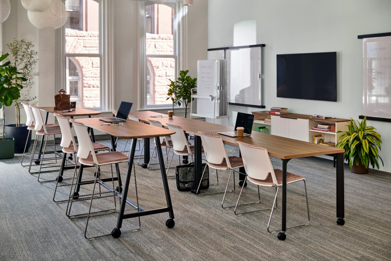 A modern conference room with a long wooden table, white chairs, two laptops, coffee cups, and large windows with natural light. The room has potted plants, whiteboards, and a large TV on the wall.