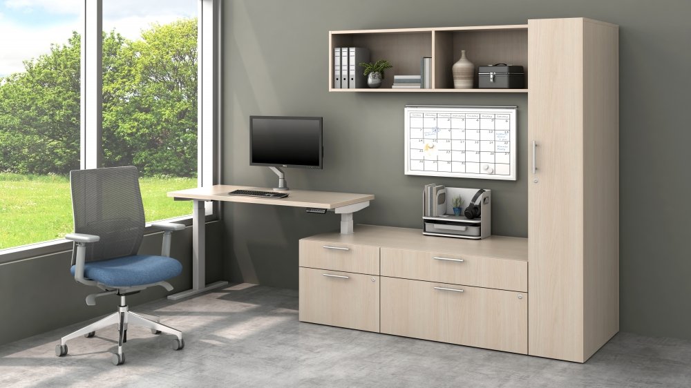 Office with a desk, gray office chair, computer monitor, wall calendar, bookshelf, and large window showing green trees outside.