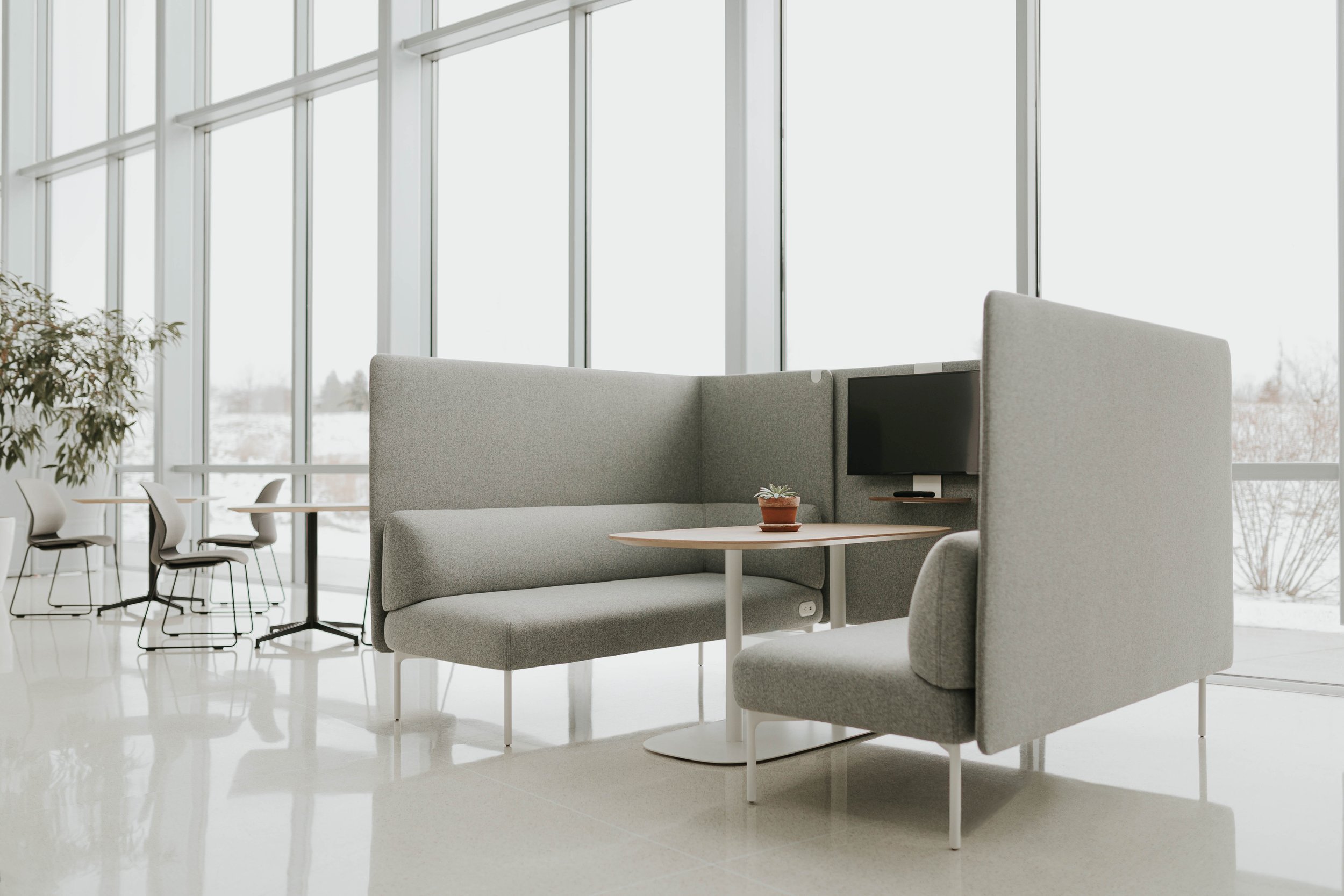 Modern office lounge area with gray seating, a small round table with a potted plant, a wall-mounted TV, large windows, and additional seating in the background.