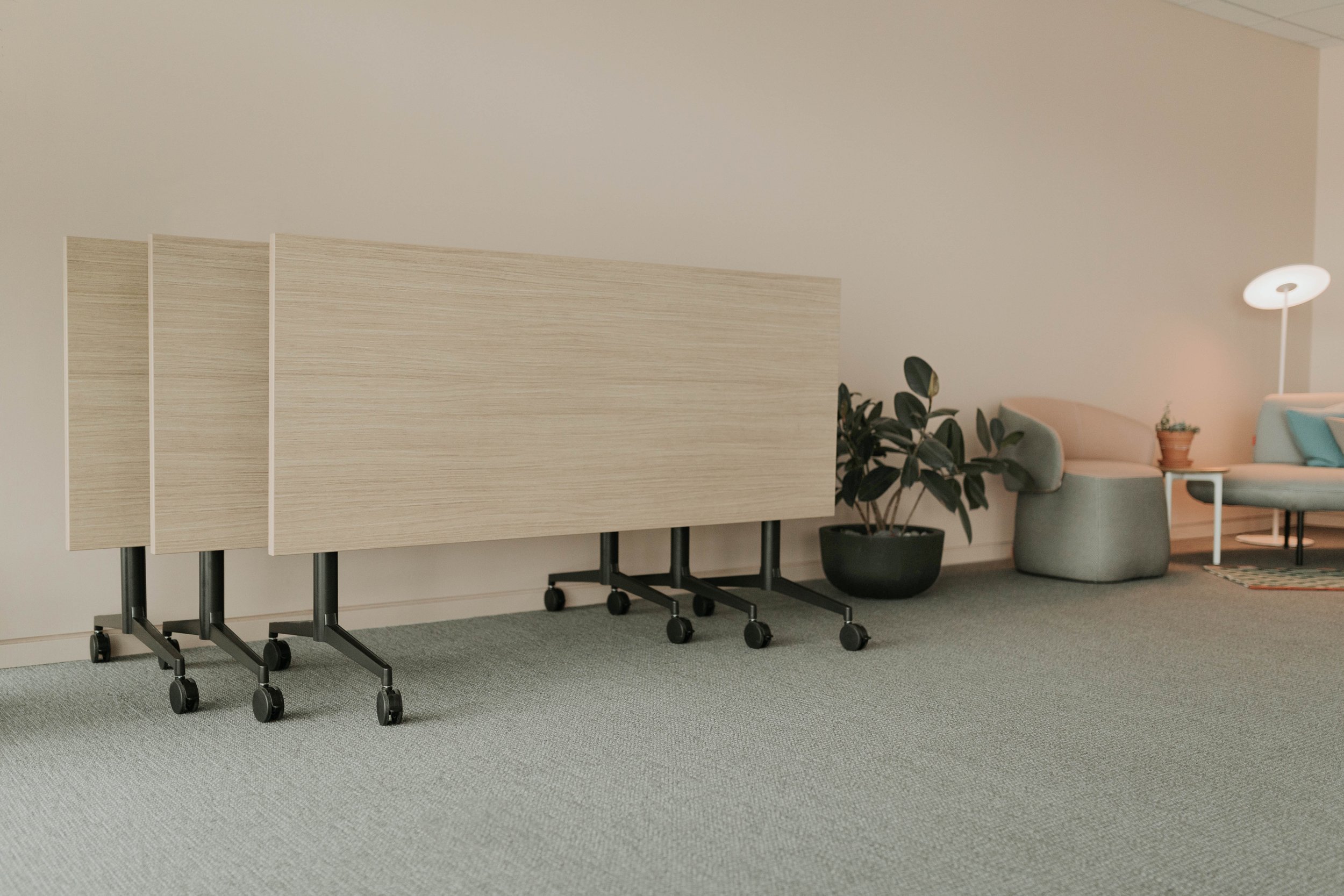 Folded beige tables on wheels next to a potted plant and a sitting area with loveseat, armchair, lamp, and decorative items in a modern lounge.