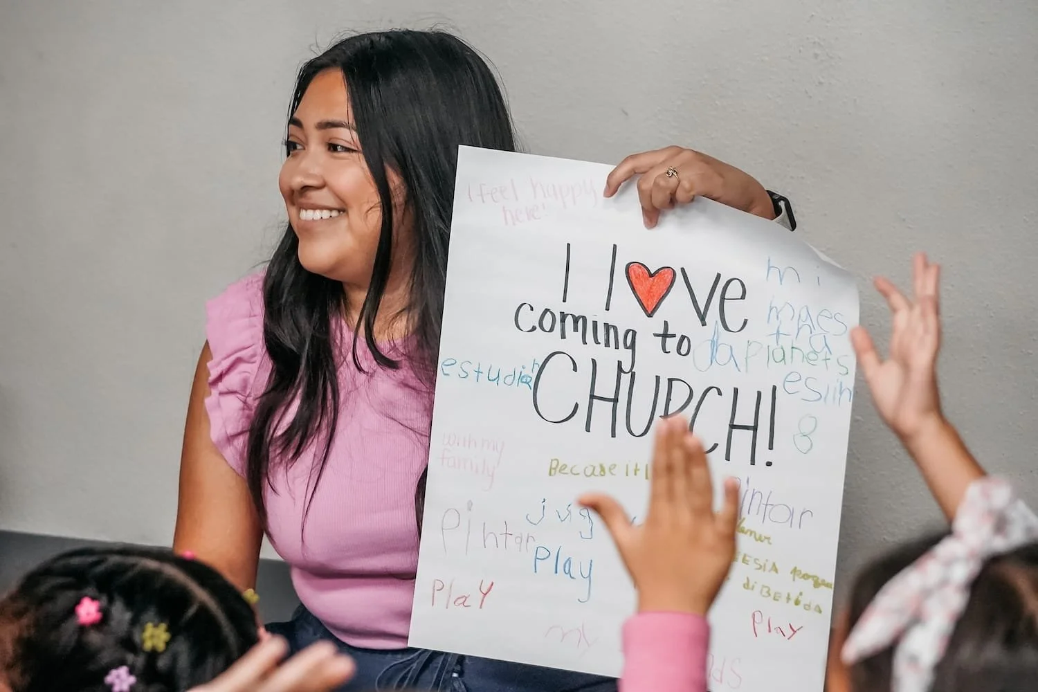 Una mujer sonriendo, sosteniendo un cartel que dice '¡Yo amo venir a la iglesia!' rodeada de niños que levantan las manos.