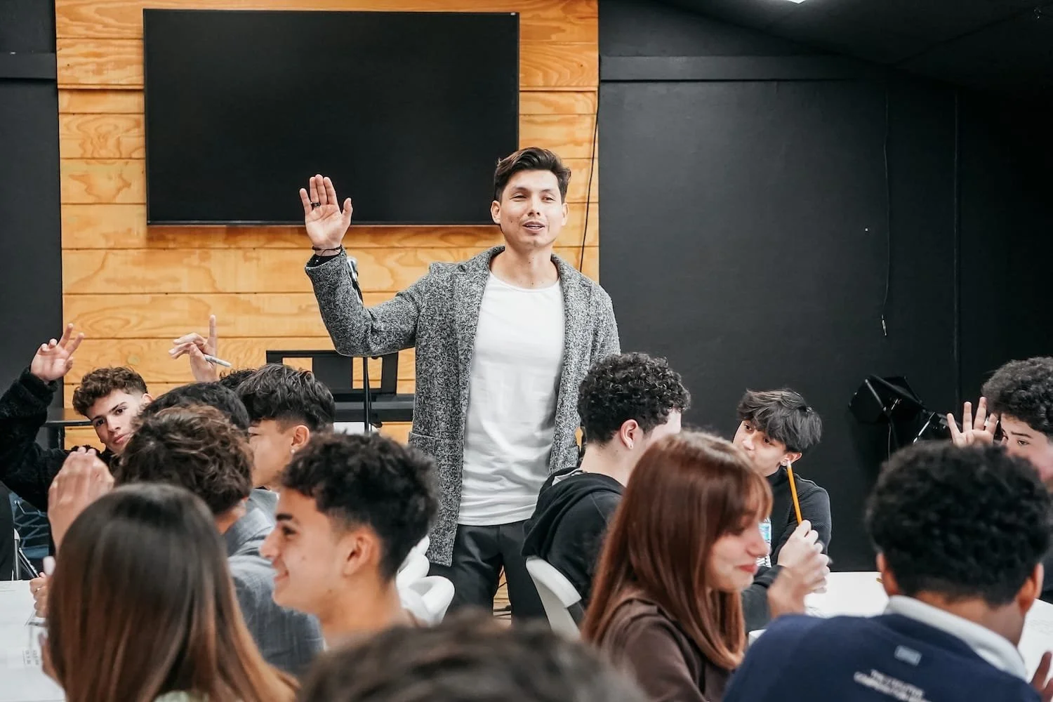 Un hombre en un salón de clases o conferencia, de cabello castaño oscuro y piel clara, con una chaqueta gris y camiseta blanca, habla y levanta la mano con expresión amable rodeado de estudiantes de diferentes etnias.