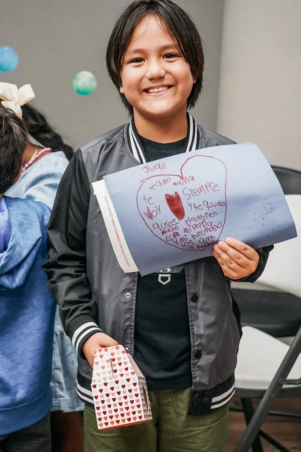 Niño sonriendo, sosteniendo una carta en forma de corazón con un mensaje escrito y un pequeño paquete con corazones de regalo.