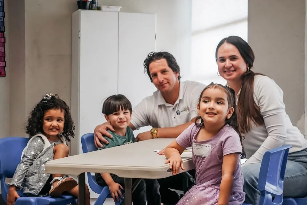 Un grupo de cinco personas, dos adultos y tres niños, sentados en una mesa en lo que parece ser una sala de clases o consulta, sonriendo a la cámara.