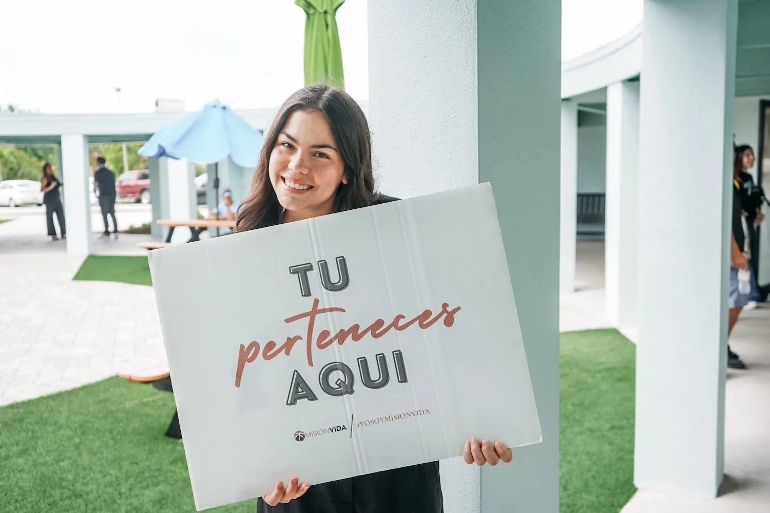 Mujer sonriendo sostiene cartel que dice 'Tú perteneces aquí' en un espacio exterior con personas y muebles en el fondo.