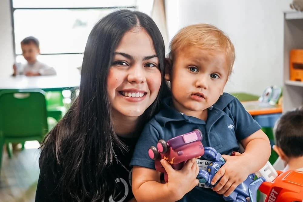 Una joven sonriente con un niño pequeño en un entorno de salón de clases con muebles verdes y otros niños al fondo.