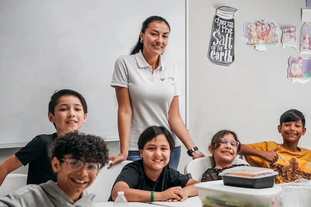 Un grupo de profesores y alumnos en un aula escolar, junto a una pizarra blanca, algunos con sonrisas y otros con expresiones relajadas.