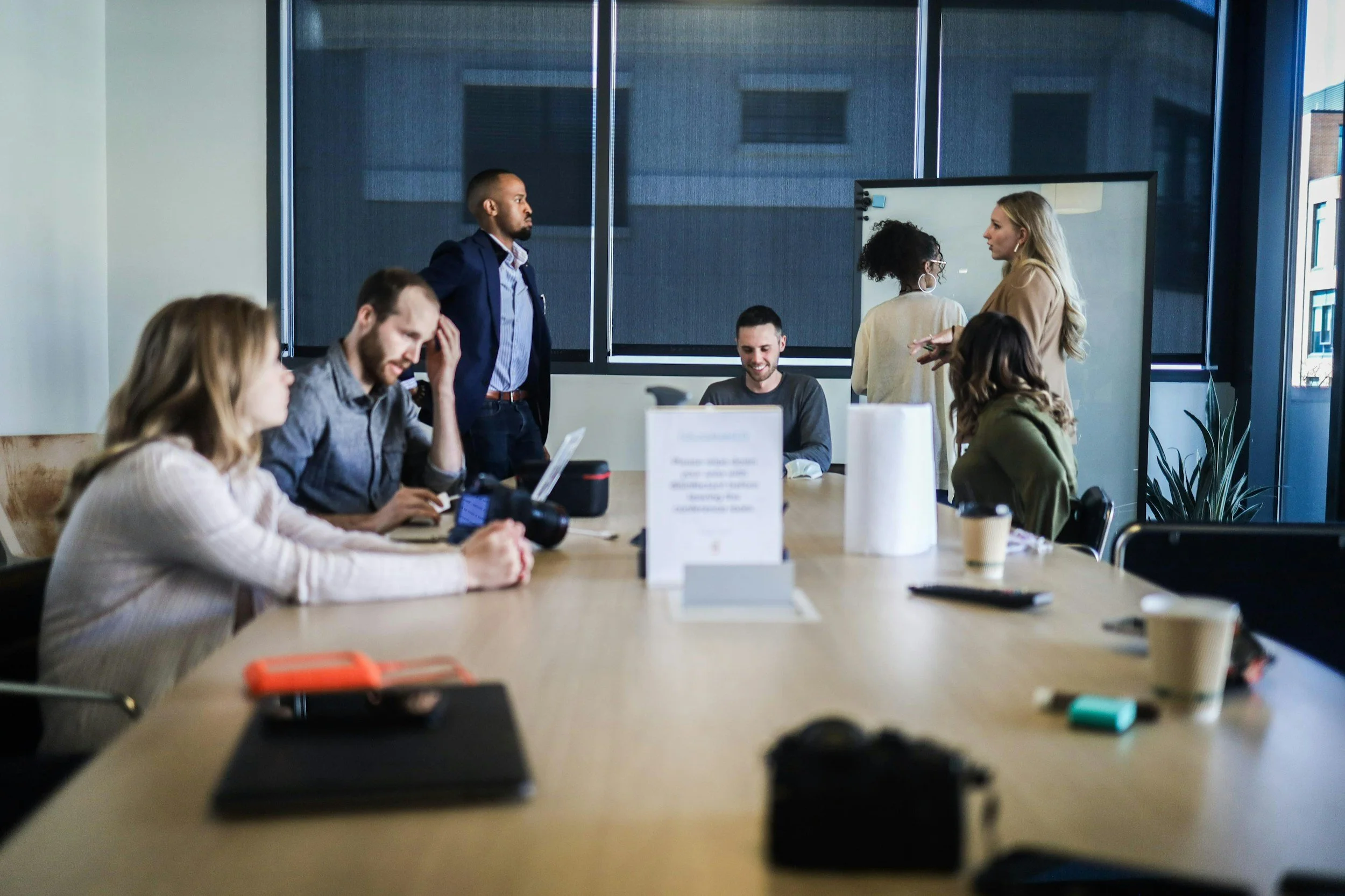 A diverse group of people in a meeting room with large windows, some sitting at a long table with laptops, coffee cups, and cameras, while others stand and converse near a whiteboard.