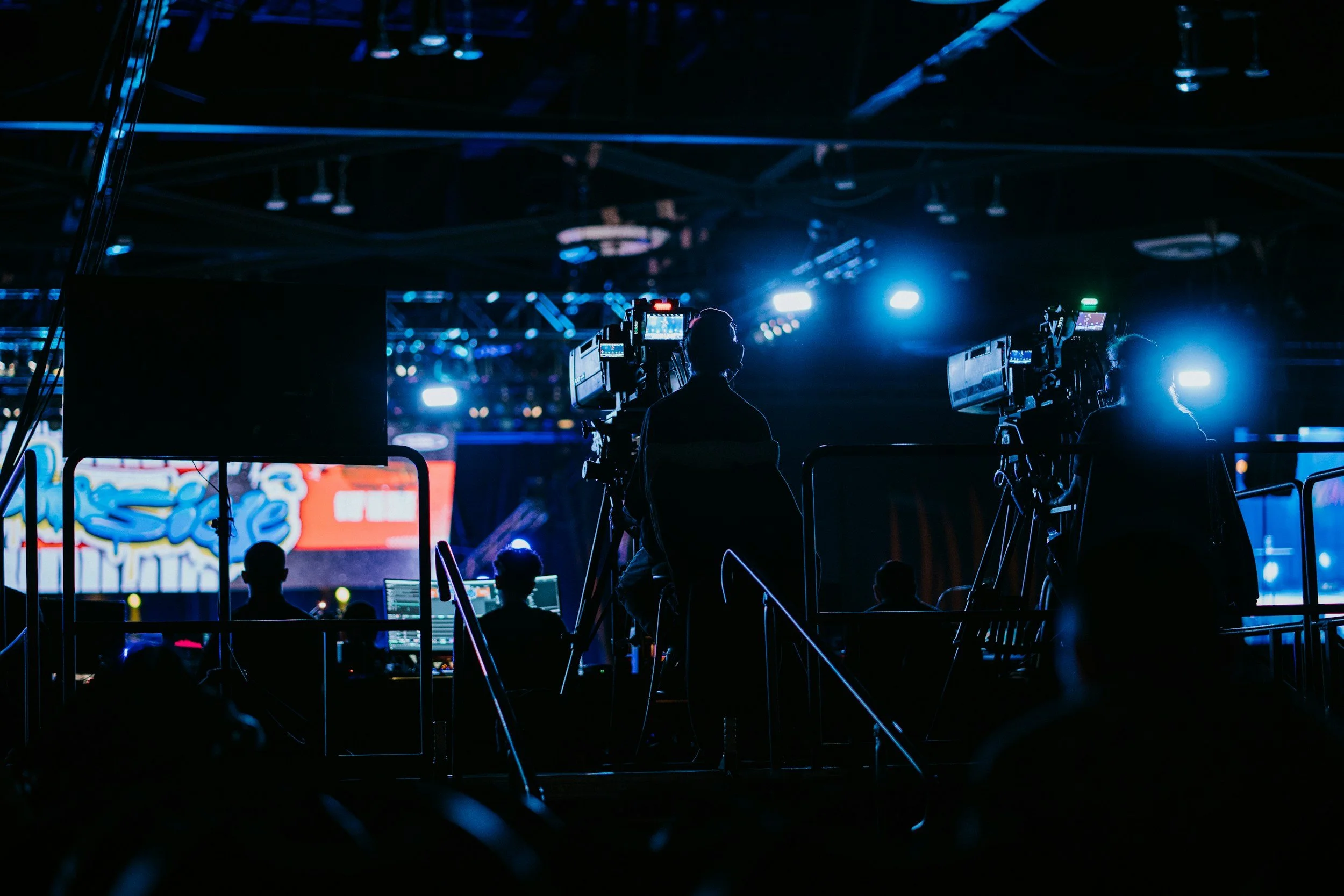 Silhouettes of camera operators and crew filming on a stage with bright blue and white stage lights, with a large screen displaying colorful graphics in the background.