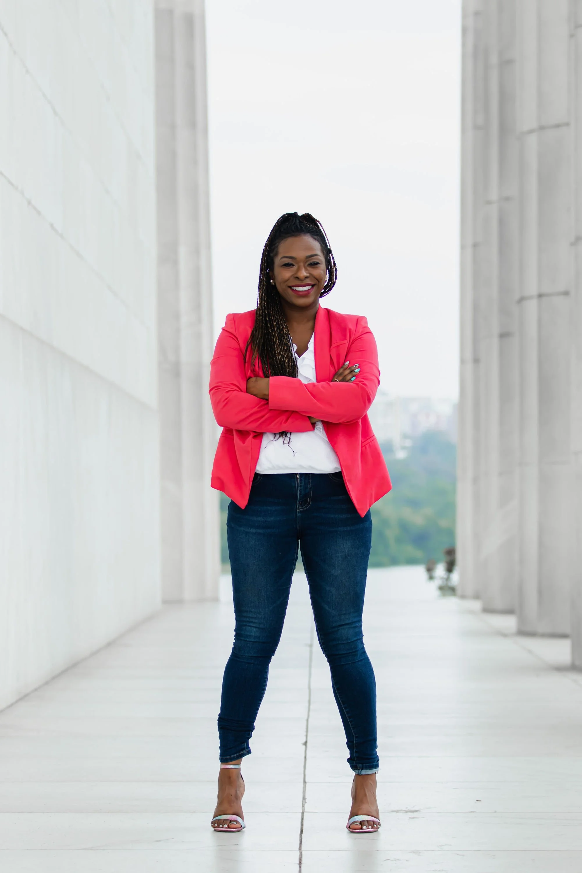 A confident woman in a coral blazer, white top, dark jeans, and white heels stands with arms crossed in an outdoor architectural setting with tall concrete columns.