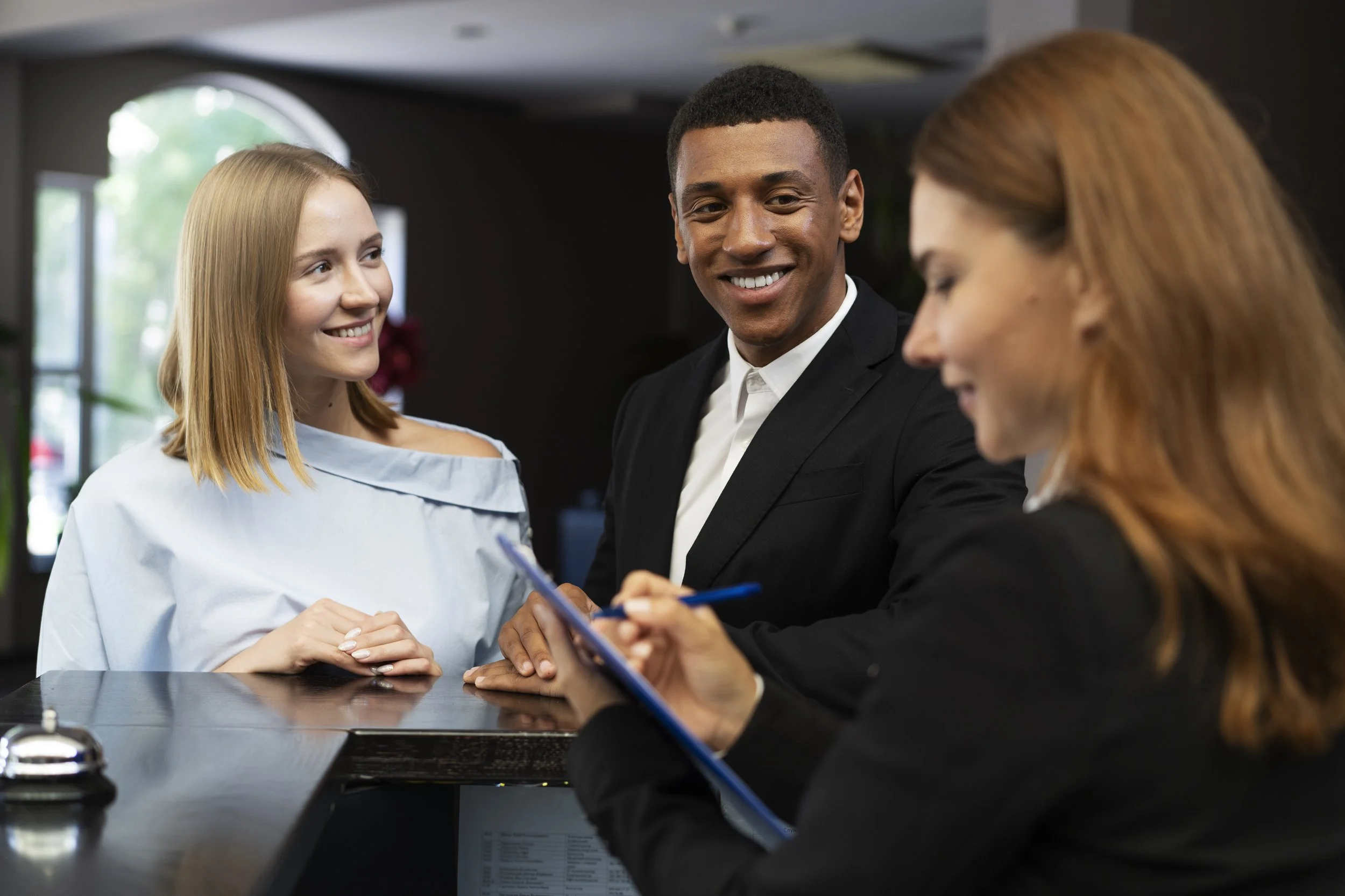 A woman at a reception desk checking in, a smiling man in a black suit talking to a woman receptionist with red hair taking notes.