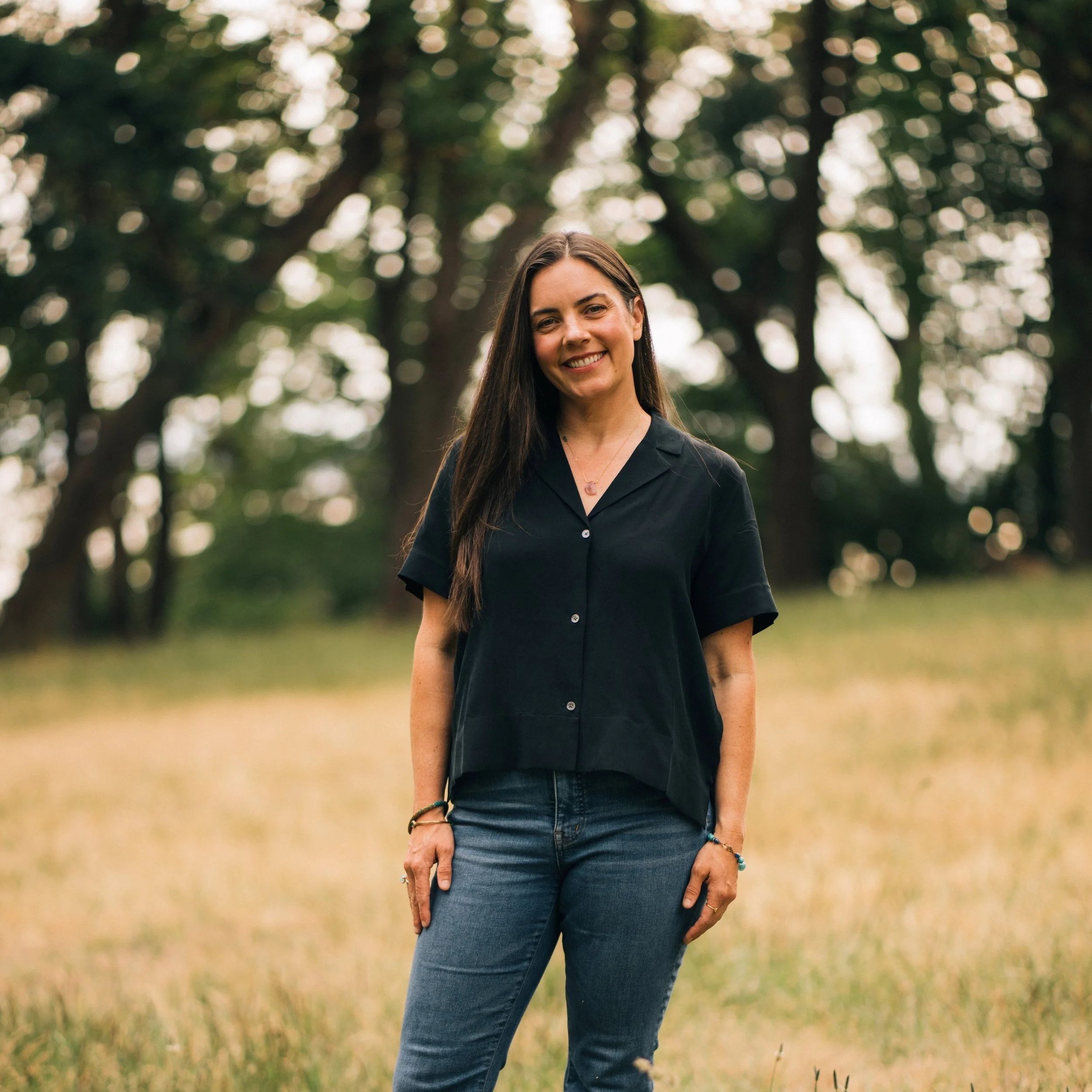 A woman with long dark hair smiling and standing in a park with trees and grass, wearing a black shirt and jeans.