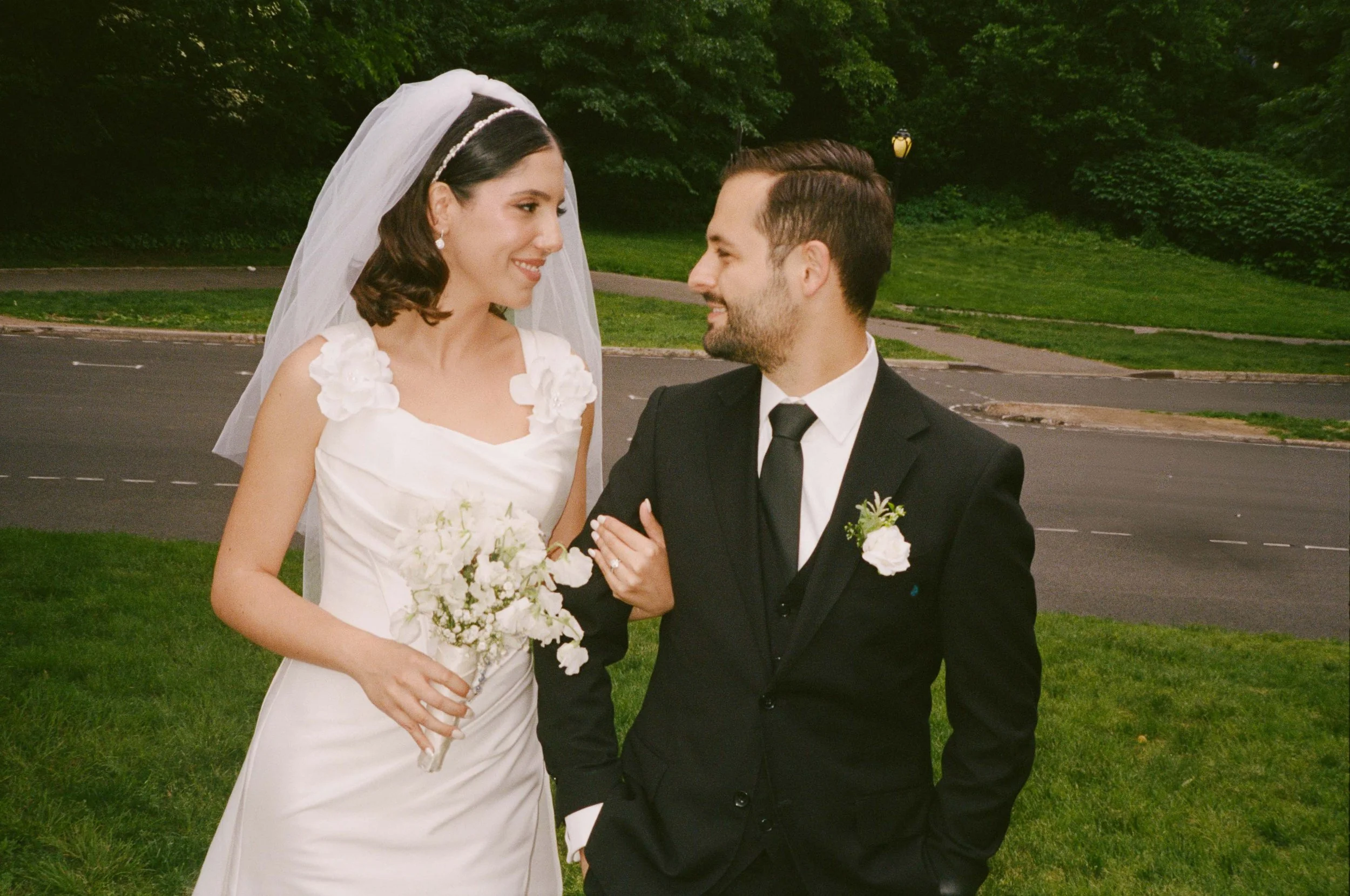 A bride and groom in wedding attire standing on a grassy area outdoors, looking at each other and smiling, with a park and road in the background.