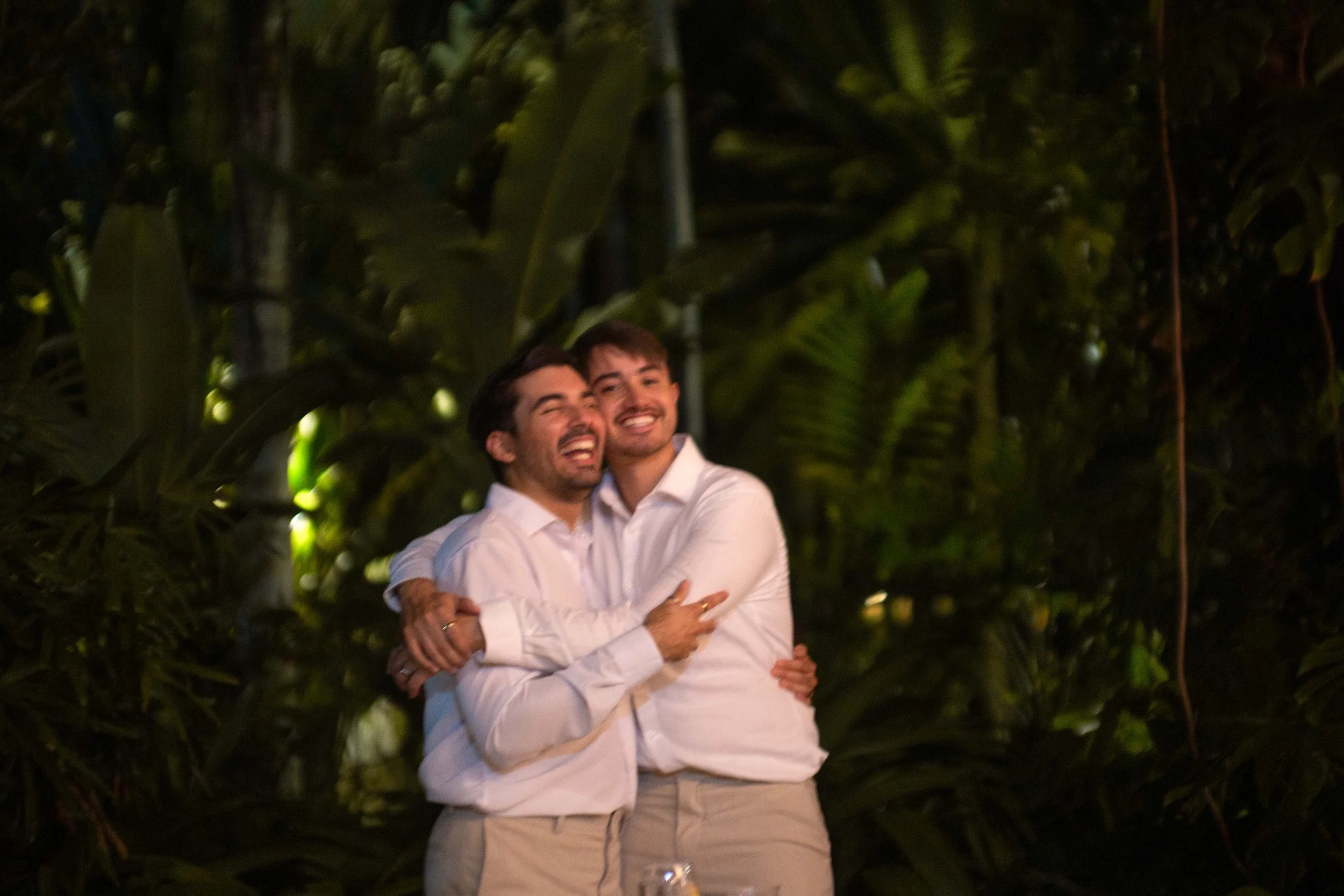 Two men wearing white shirts hugging and smiling, standing outdoors at night surrounded by lush green tropical foliage.