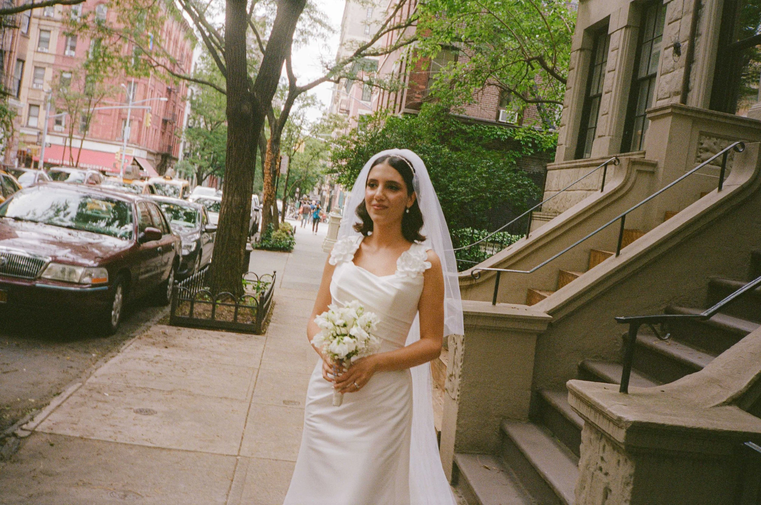 A bride standing on a city sidewalk holding a bouquet of white flowers, wearing a white wedding dress with floral details on the shoulders and a veil.