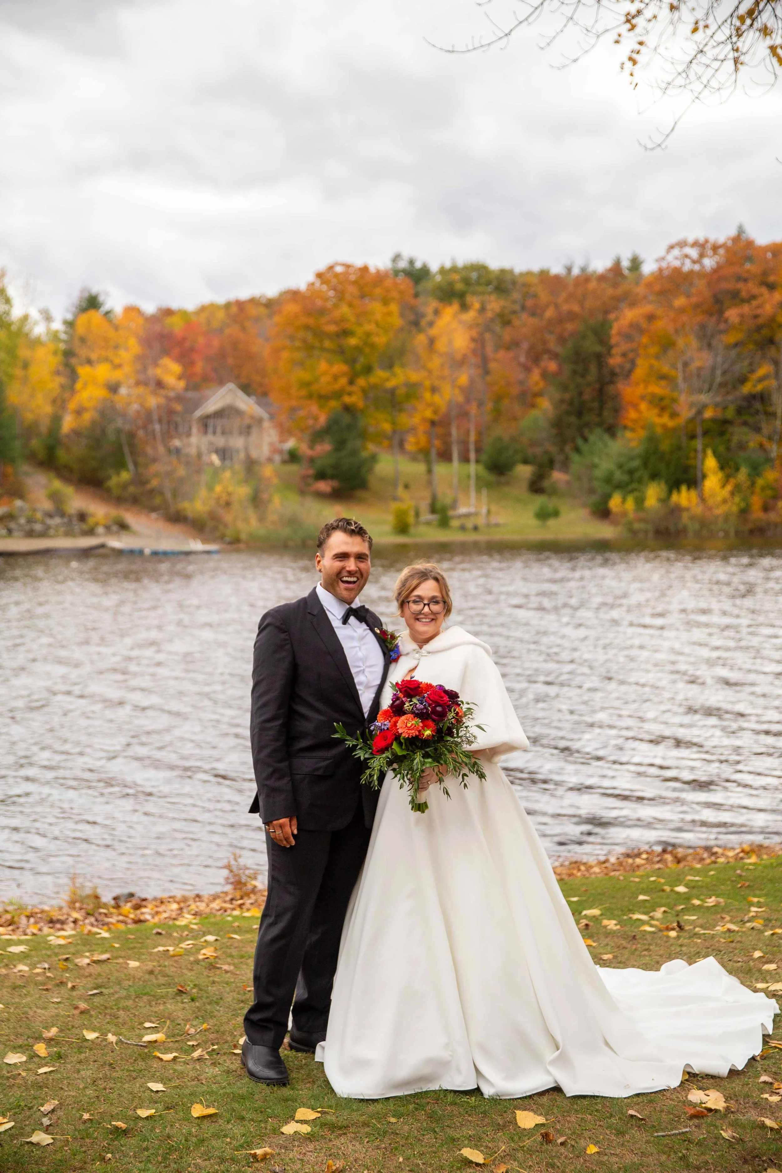 A newlywed couple in wedding attire standing outdoors by a lake with autumn-colored trees in the background, smiling at the camera.