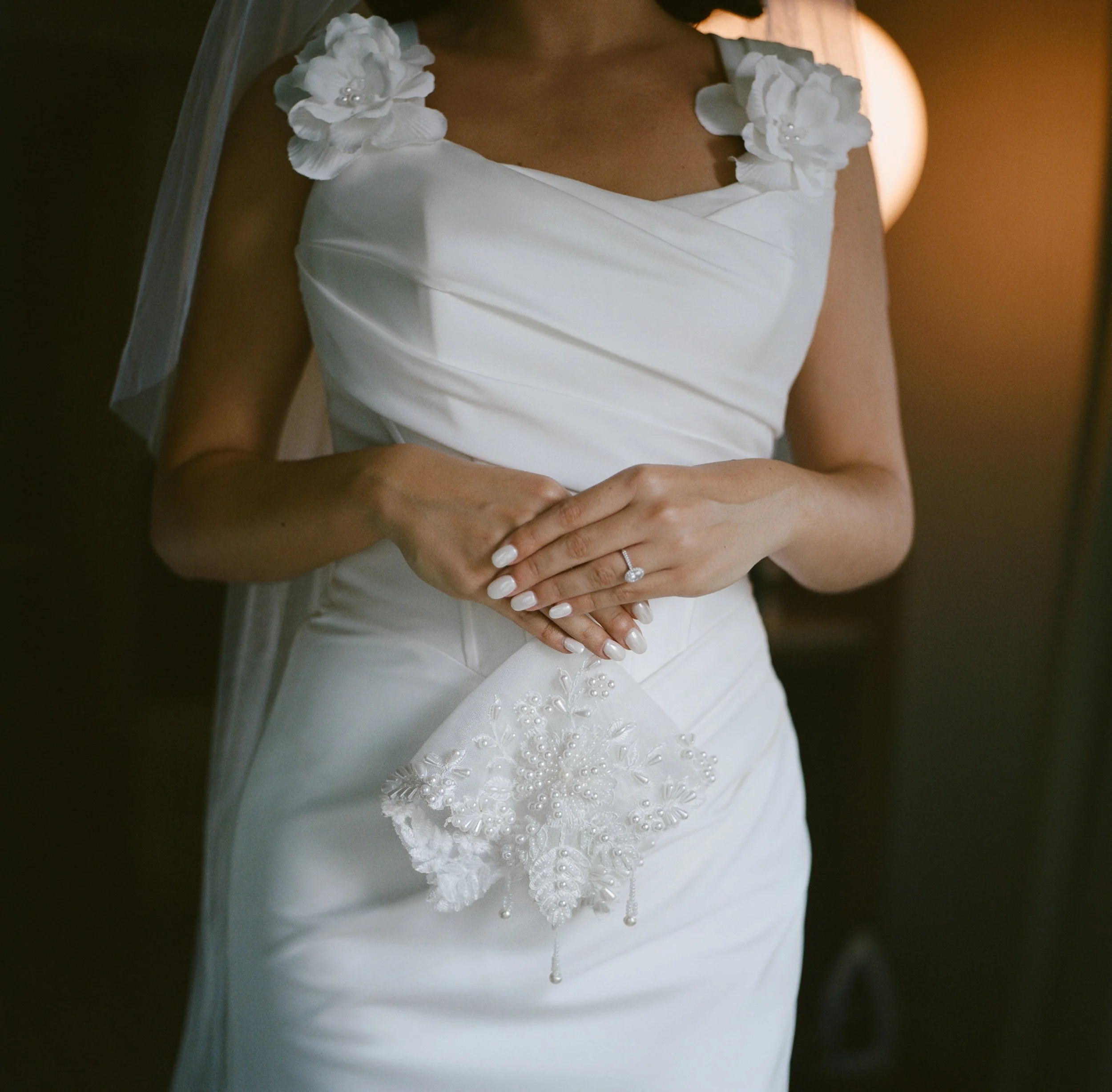 A woman in a white wedding dress holding a pearl-embellished clutch purse, with her hands clasped, showcasing a diamond ring. She is also wearing floral detail on her shoulders.