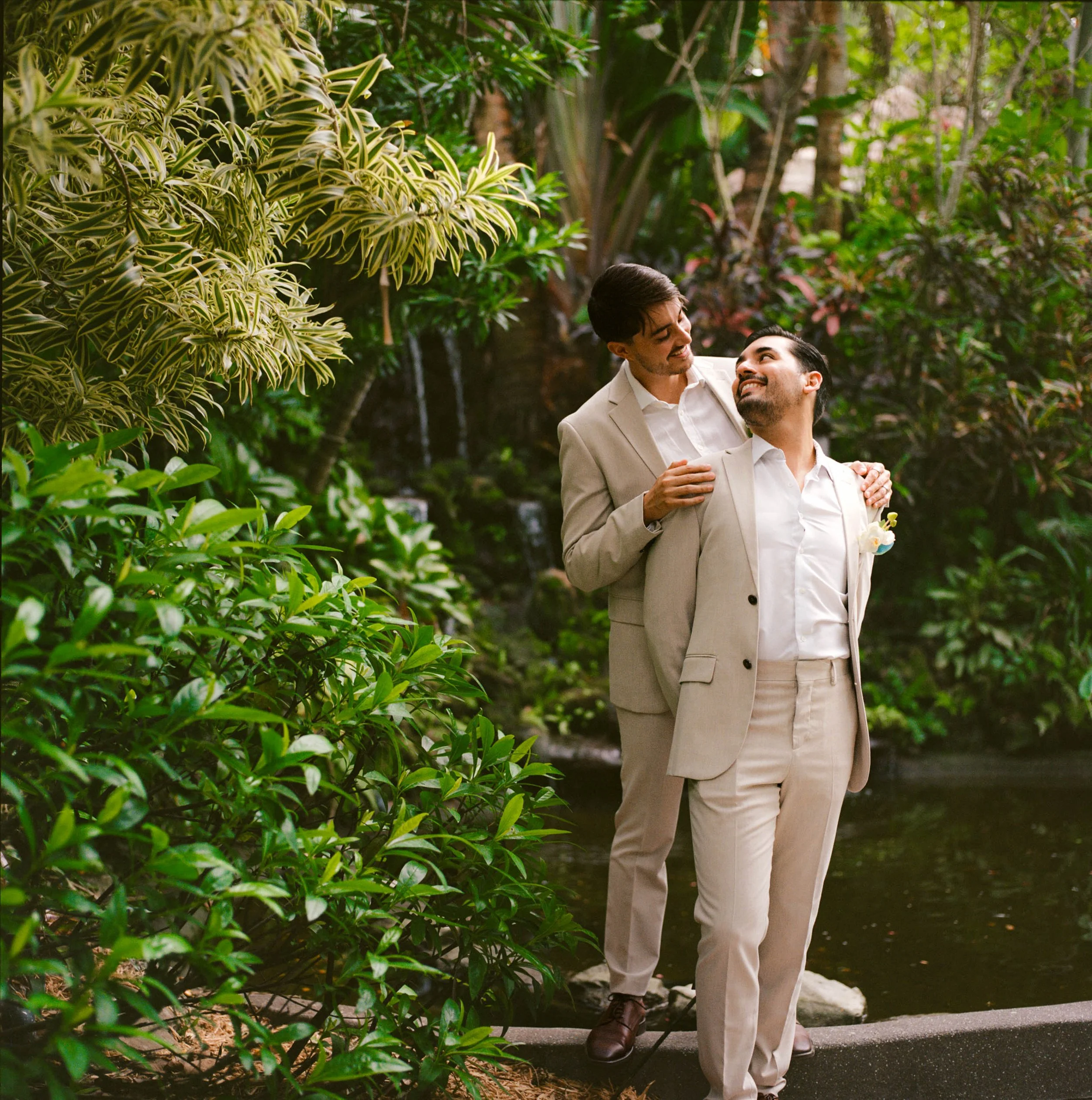 Two men in light-colored suits standing close together in a lush garden, smiling and looking at each other, with greenery and a small waterfall in the background.