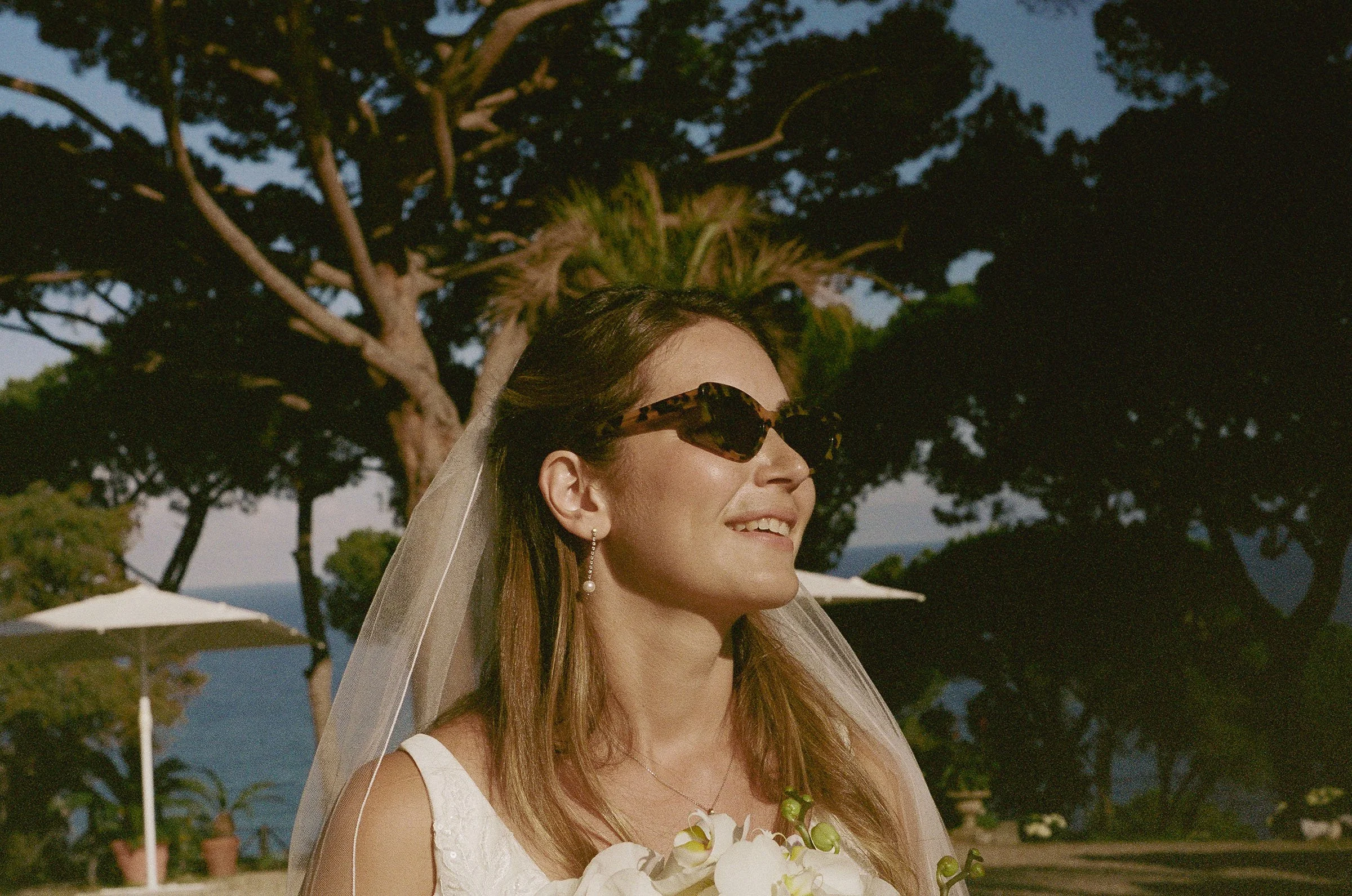 A woman in a wedding dress with a veil and sunglasses, smiling outdoors during the day, with trees and umbrellas in the background.