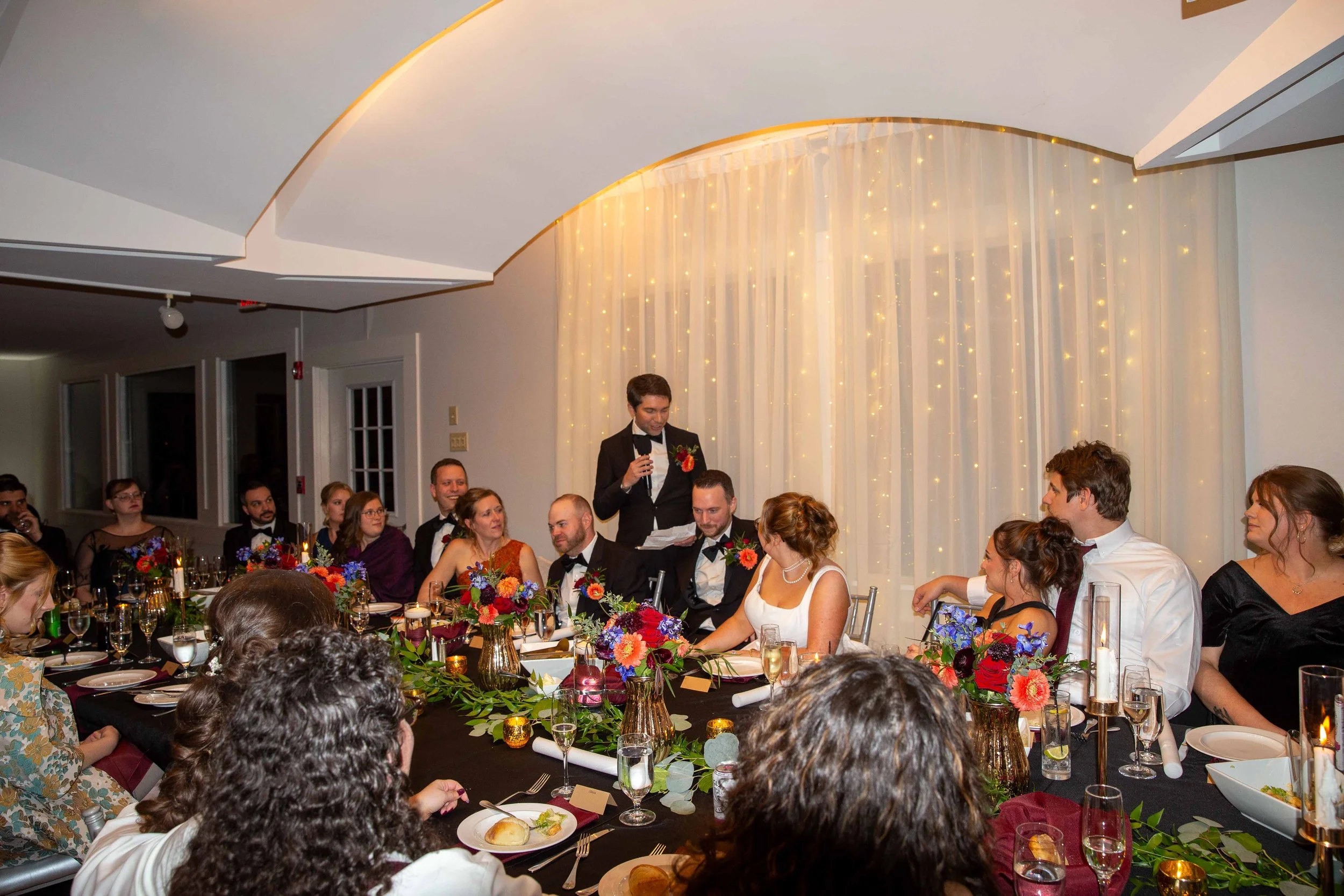 A wedding reception with guests seated at a long table, a man giving a toast, and a backdrop of fairy lights and curtains.