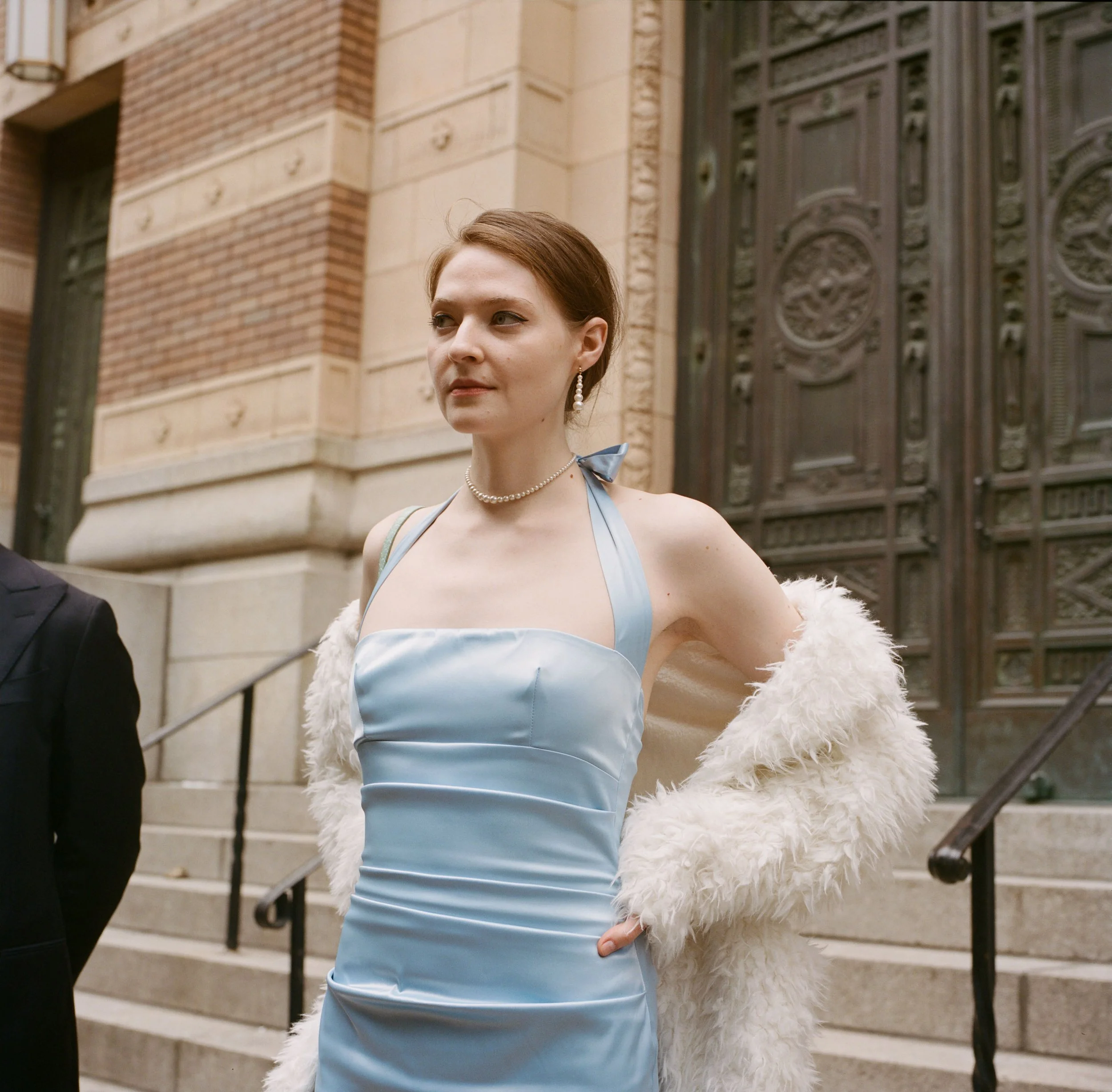 A woman in a light blue satin dress with a halter strap, pearl necklace, and pearl earrings, standing on steps outside a brick and stone building with ornate metal doors, wearing a white fluffy faux fur coat draped over her shoulders.