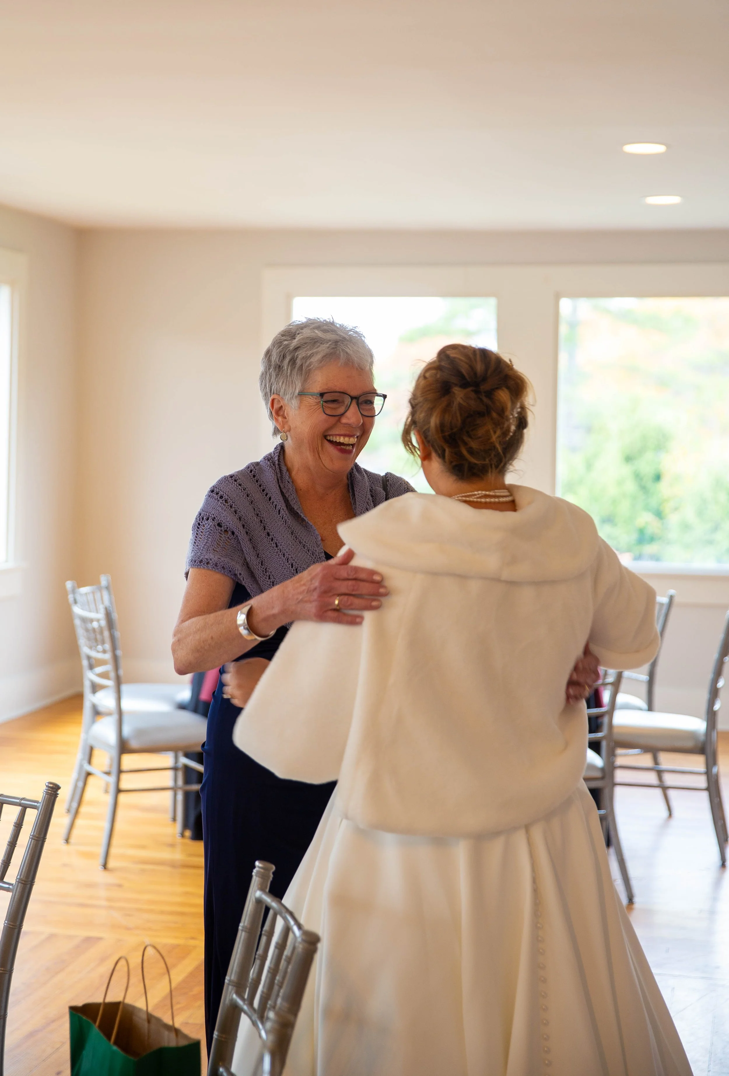 An elderly woman and a bride sharing a joyful embrace in a bright, decorated room with chairs and large windows.