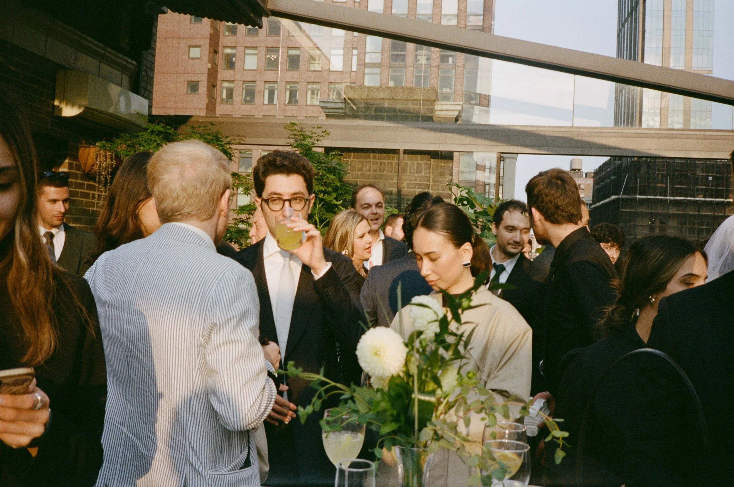 A group of people at a social gathering or cocktail party on a rooftop terrace in an urban setting, with high-rise buildings in the background. People are dressed in formal and semi-formal attire, some holding drinks, and there is a floral centerpiec