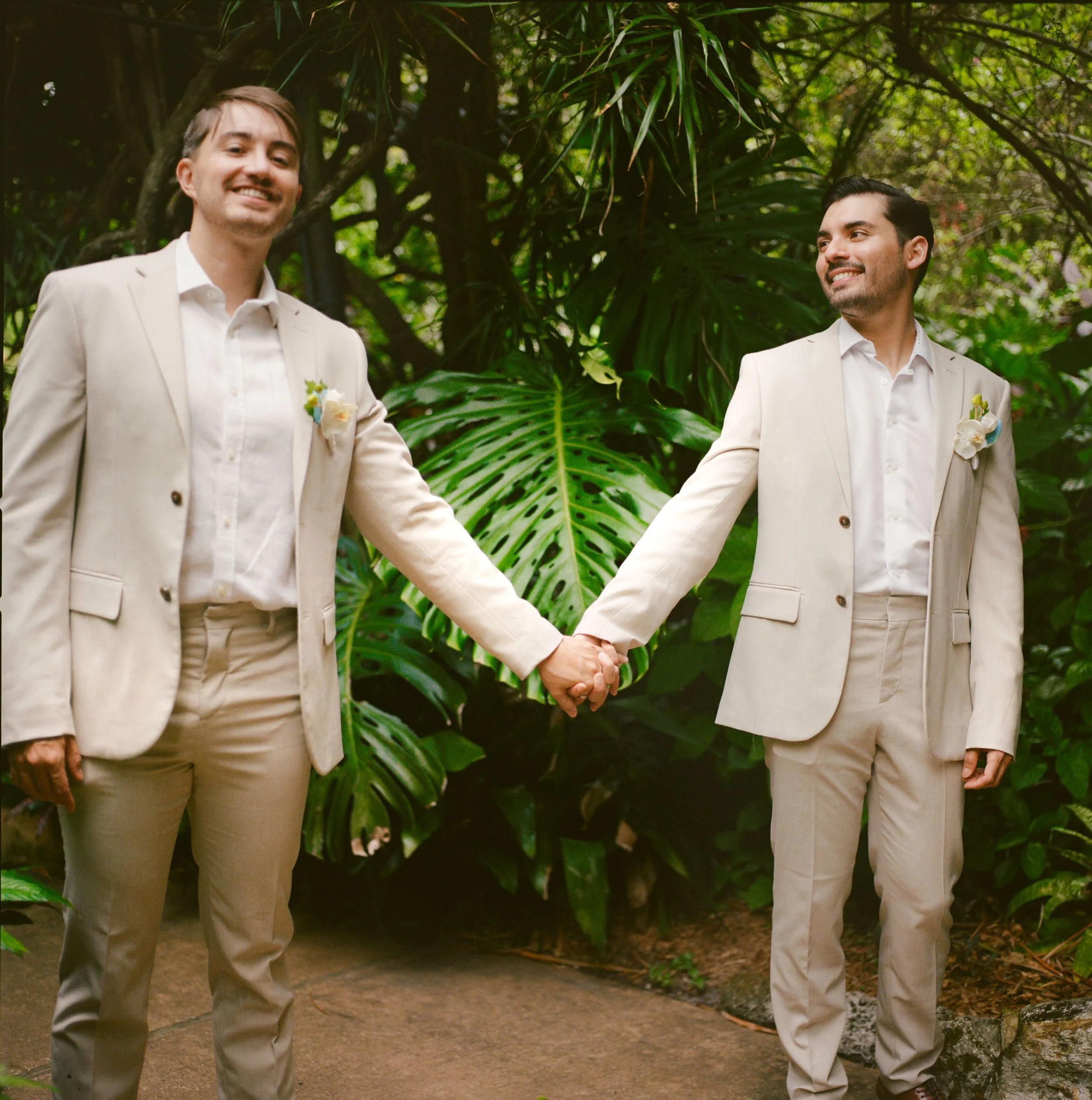 Two men in cream-colored suits holding hands in a lush green garden.
