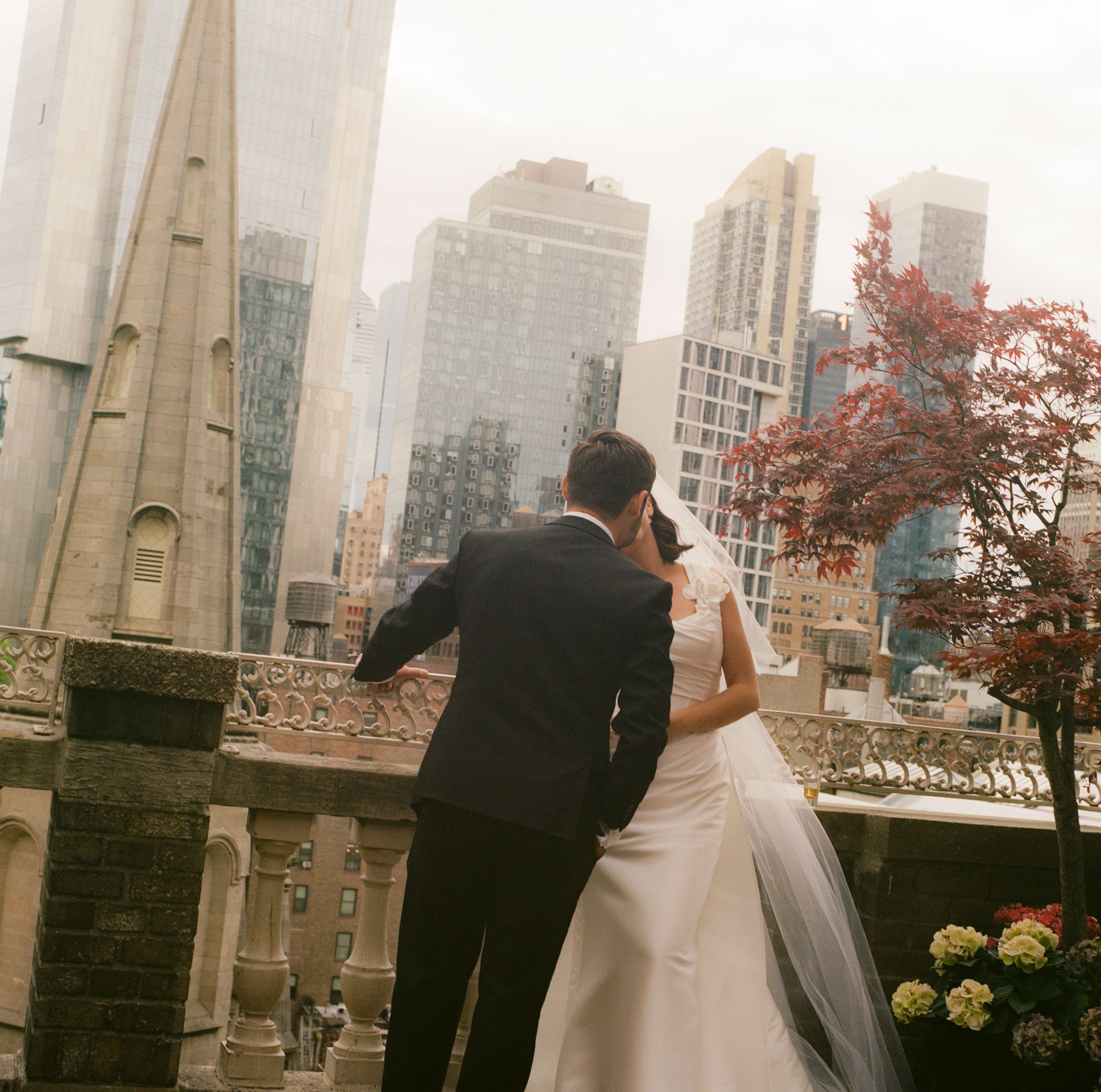 A bride and groom sharing a kiss on a balcony rooftop with New York City skyscrapers in the background, some with a tree and flowers nearby.