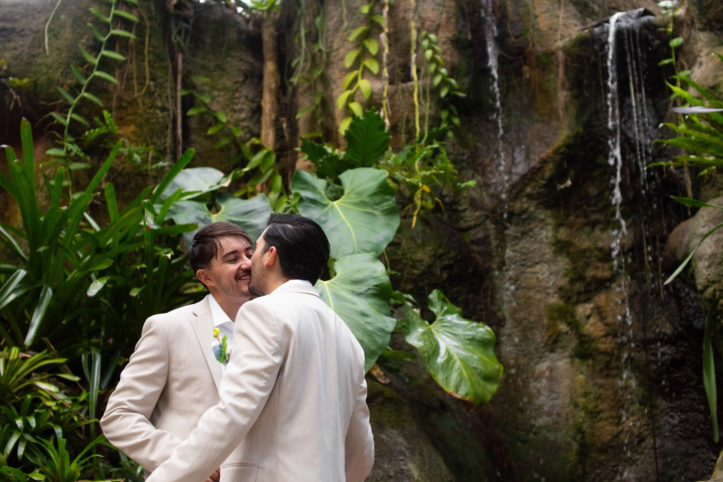 Two men in white suits sharing a kiss in front of a lush, tropical backdrop with large green leaves and a small waterfall.